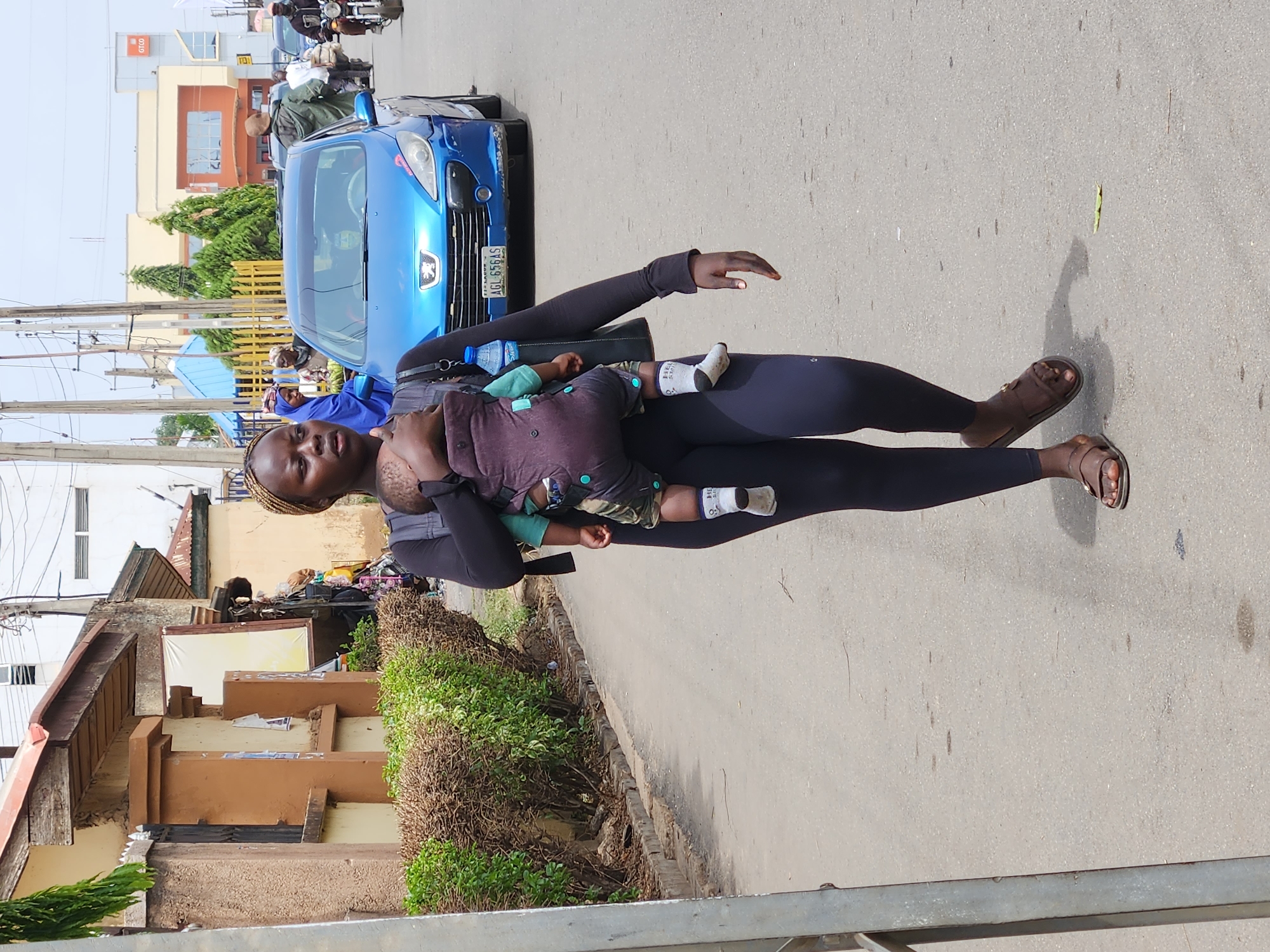 A woman carries a baby in a carrier while standing on a paved street in Jos, Nigeria. The woman is wearing a long-sleeved black shirt and leggings, and brown sandals. The baby is wearing a green shirt, patterned shorts, and a white sock on one foot. Behind the woman, a blue car is parked on the side of the street. Buildings and greenery are visible in the background. A motorcycle with two riders is visible in the background.