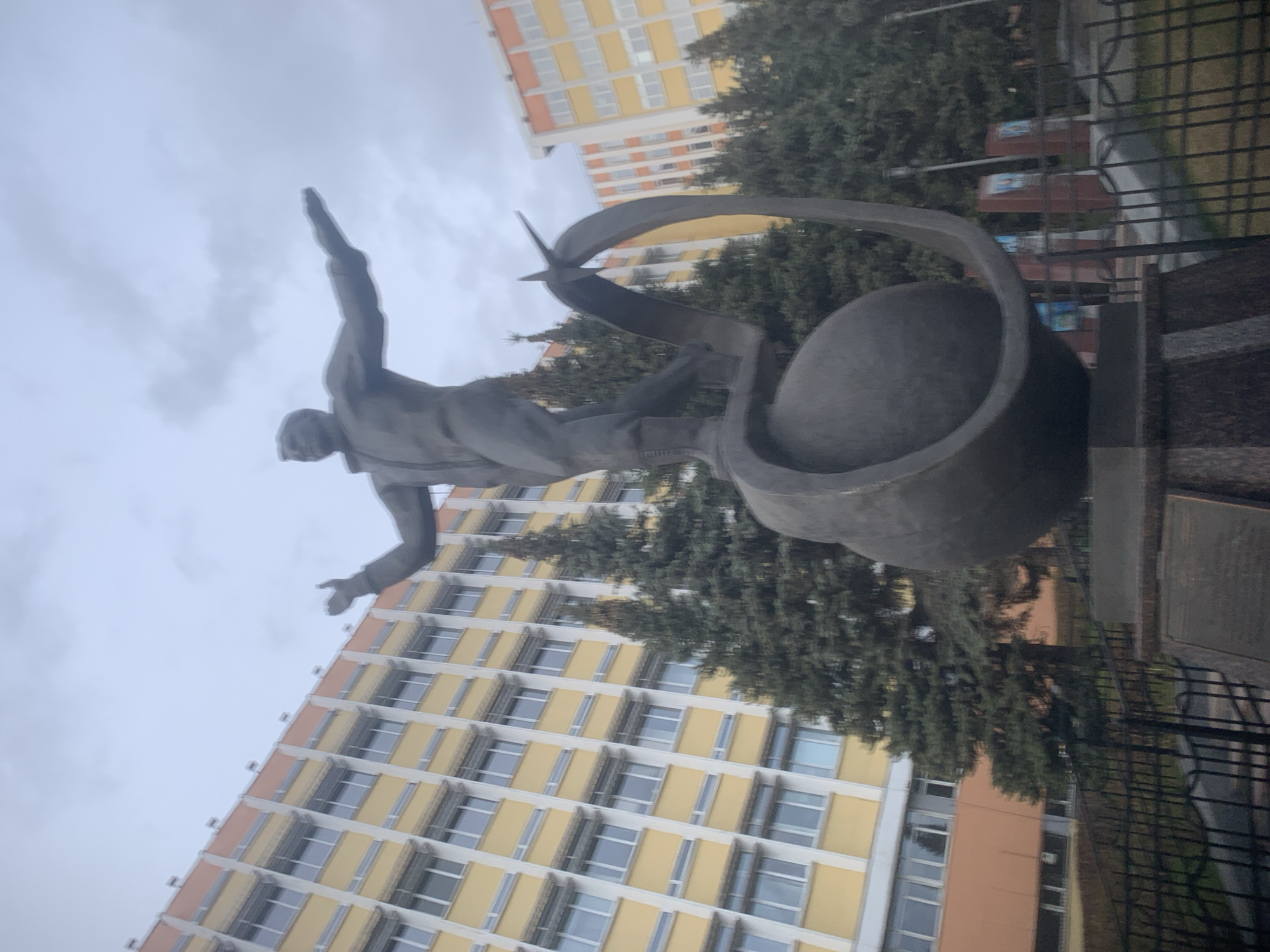 The image displays a statue dedicated to flight or space exploration, set against an urban backdrop in Izhevsk, Russia. The central subject is a dark-colored, likely bronze or stone, statue of a male figure in a dynamic pose. The figure appears to be leaping or flying upwards, with arms outstretched—one pointing directly skyward and the other slightly to the side. His posture suggests forward motion and aspiration.
Behind and around the figure is an abstract, curving element resembling an orbital path or a swooping ribbon, which encircles a spherical object, possibly symbolizing Earth or a celestial body. A small, stylized airplane or rocket is integrated into this abstract design, positioned behind the figure's head, reinforcing the theme of aviation or space travel.
In the background, a large, multi-story building with yellow and orange facades is prominent, indicating an urban environment. Several tall, dark green coniferous trees are interspersed between the statue and the buildings, adding a natural element to the scene. The sky is uniformly light grey and overcast, suggesting a cloudy day. A black metal fence is visible along the right edge of the foreground, separating the monument area from what appears to be a pathway or street. No visible text is discernible on the statue or the surrounding elements. The overall impression is a tribute to human endeavor in the realm of flight or space within a typical city setting.