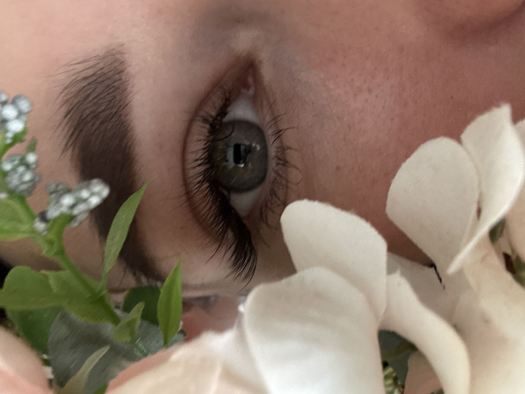 A close-up, slightly angled view captures a human eye surrounded by floral elements. The eye, featuring long eyelashes and a dark pupil, gazes upwards. Above the eye, a shaped eyebrow is visible. The surrounding environment includes white petals and green foliage, potentially artificial flowers. The skin is fair and smooth. No specific details indicate a location in Chaikovsky, Russia. The scene focuses primarily on the eye and surrounding floral arrangement.