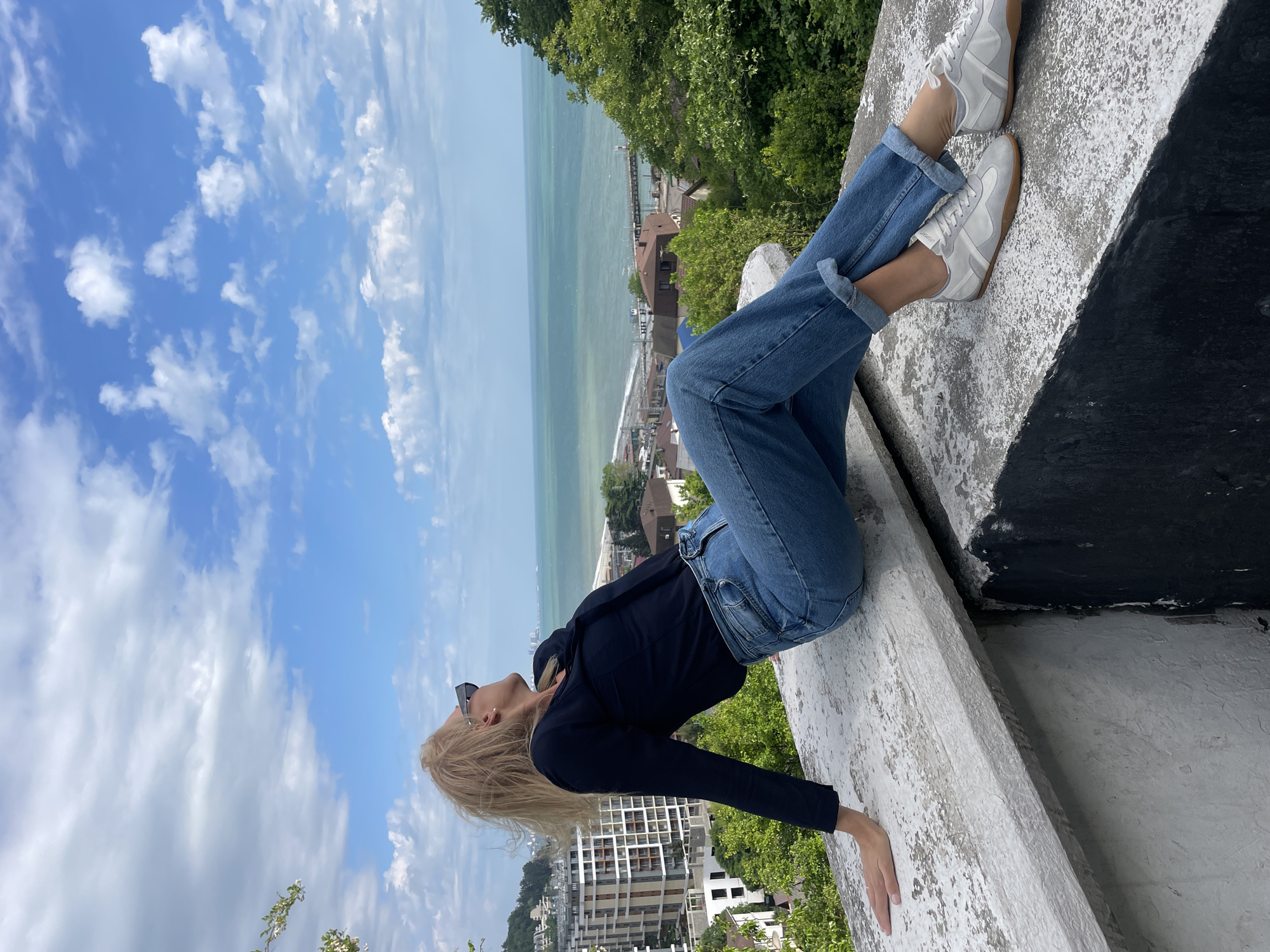 A woman is sitting on a ledge overlooking Dagomys, Russia. She is wearing blue jeans, a black jacket, and white sneakers. She is leaning back and looking up at the sky. The sky is blue with white clouds. In the distance, there is a view of the sea and the city. The woman's right hand is resting on the ledge.