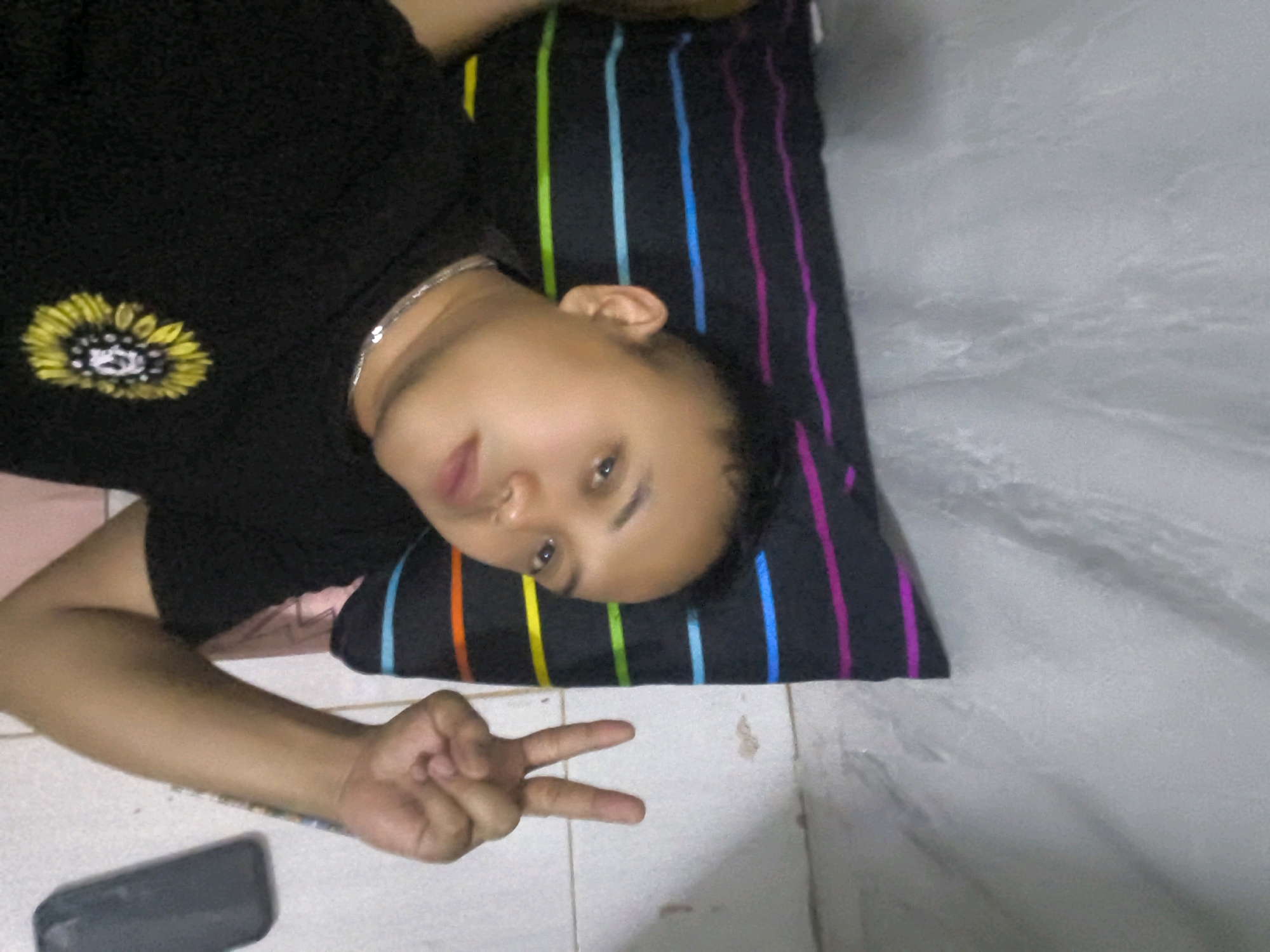 A person lies on a striped pillow on a tiled floor, with their head to the right. They are wearing a black t-shirt with a sunflower emblem. Their left arm is visible, with a hand held up making a peace sign. A dark rectangular object, possibly a phone, is on the floor to the left of the person. The pillow has vertical stripes of various colors including green, yellow, blue, and pink. The background is a light-colored tiled surface. The lighting is dim, and the image is slightly blurry.