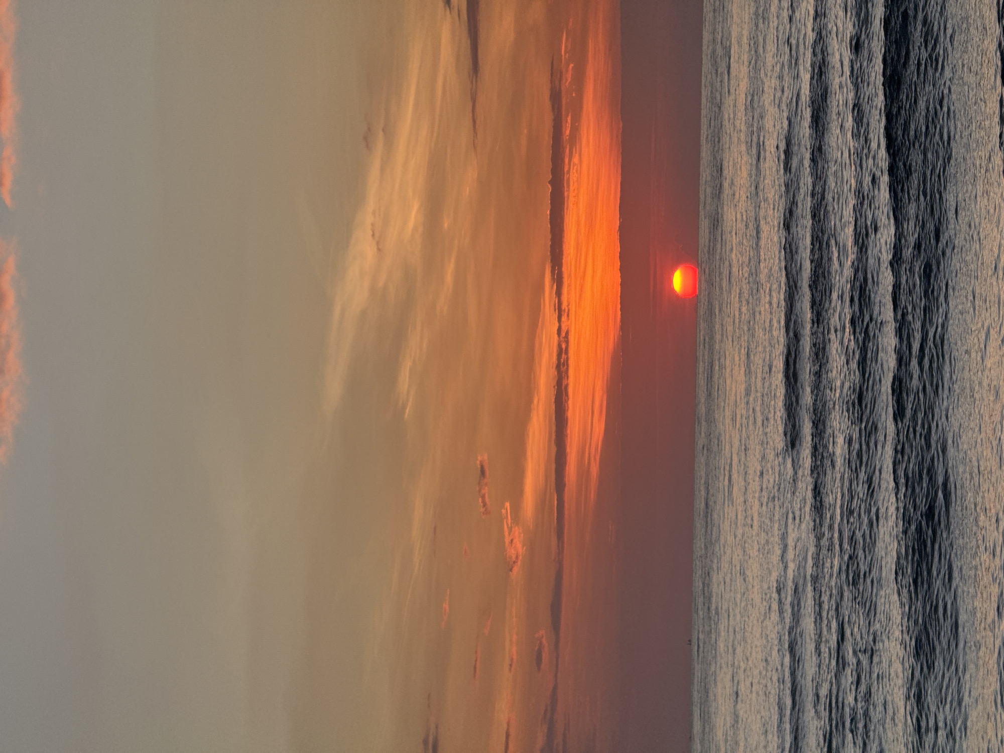 The image depicts a serene seascape with a prominent sunset. The sky is painted with a gradient of colors, transitioning from a pale, grayish-peach at the top to vibrant oranges and reds closer to the horizon. Wispy clouds are scattered across the sky, catching the warm hues of the setting sun. The sun itself is a brilliant, fiery orb, partially submerged below the horizon, casting a warm glow on the surrounding atmosphere.
The ocean stretches out in front, characterized by gentle waves that create a textured surface. The water reflects the colors of the sky, with darker blues and grays dominating the foreground, gradually lightening towards the horizon where it meets the fiery sky. There are no discernible people or structures in the immediate foreground or mid-ground. In the far distance, on the horizon, a very small, dark silhouette that could be a boat or a distant landmass is faintly visible.
The scene is tranquil and evokes a sense of natural beauty and calm. The image's composition, with the vast expanse of the sky and ocean, suggests an open, natural environment. Given the provided context, this seascape is located in the city of Abang, Indonesia. The specific location within Abang would be a coastal area, likely a beach or a vantage point overlooking the ocean.