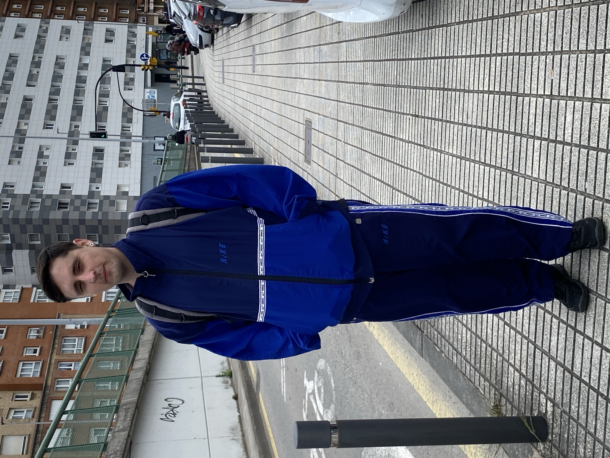 A man stands on a paved sidewalk in Gijón/Xixón, Spain. He wears a blue Nike tracksuit and black shoes. The sidewalk is made of rectangular gray stones laid in a grid pattern. Behind the man are buildings of varying heights and architectural styles. A street runs parallel to the sidewalk, with parked cars visible. Traffic lights are positioned along the street. A gray metal railing and a concrete wall with graffiti are visible on the left side of the frame. A dark, cylindrical metal bollard stands in the foreground.