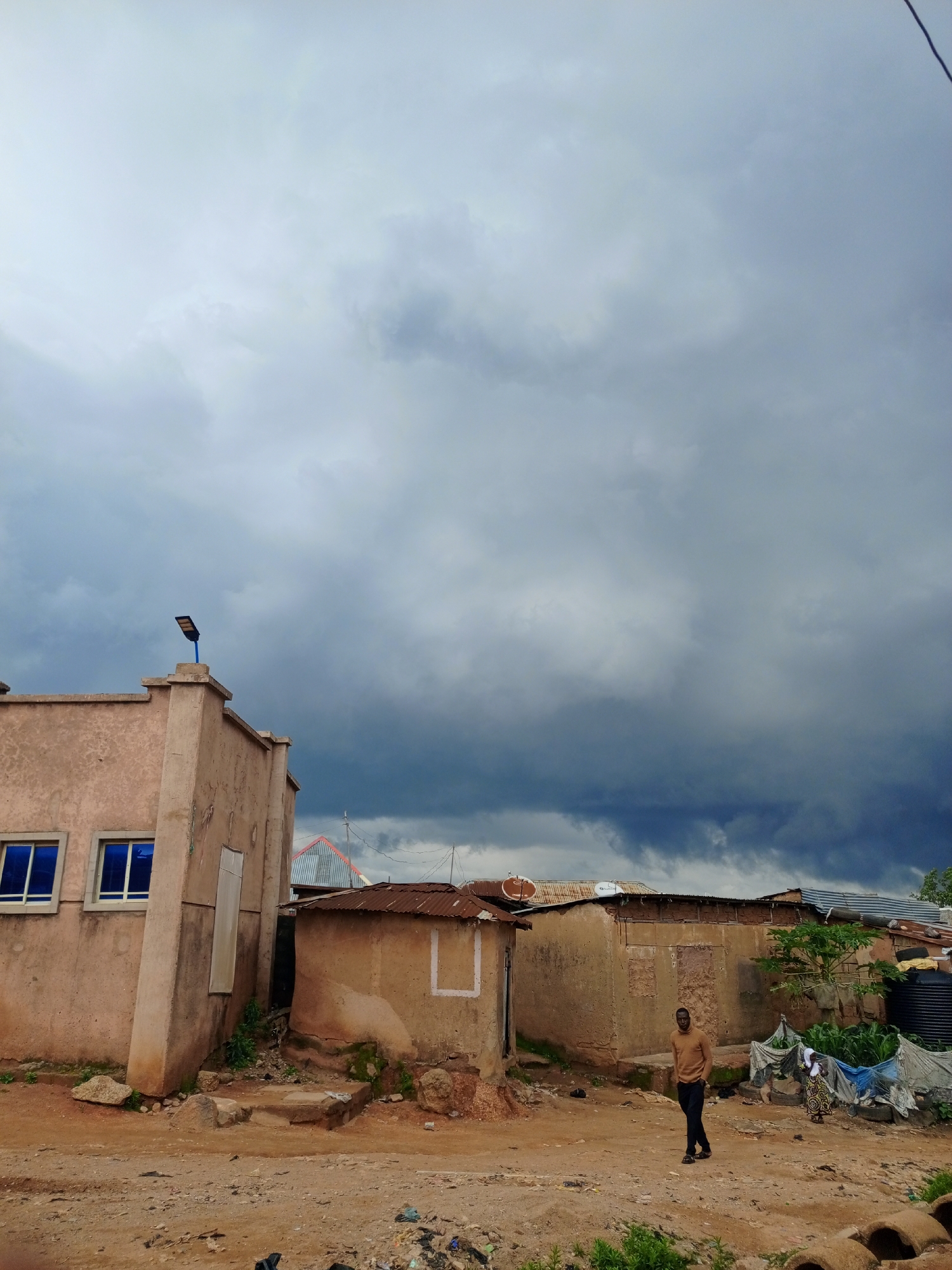 In the city of Jos, Nigeria, a street scene unfolds under a dramatic, overcast sky filled with dark, looming clouds, suggesting an impending storm. The foreground is a dusty, unpaved road, littered with debris including plastic bags, and what appear to be discarded ceramic pipes or rings. To the left, a two-story building with a light-colored, rough-textured facade stands prominently. It has two rectangular blue windows on its upper level and a tall, narrow white-framed window or door on its lower level. A modern blue street light is mounted on the corner of its roof.
Adjacent to this building, a series of smaller, less structured dwellings stretch across the mid-ground. These structures have walls made of earth or concrete, with corrugated metal roofs, some showing signs of rust. One building has a small, partially visible doorway with plants growing around it. Another has a white rectangular outline on its exterior wall, possibly a window or decorative element.
A man dressed in a light brown turtleneck sweater and dark pants stands in the middle of the dirt path, facing slightly to the right, appearing to walk. Further back, near the dwellings, a woman in a patterned dress and headscarf is visible, standing next to a patch of green plants covered partially by a blue tarp. Tires are stacked near the plants, and a large black water tank is situated behind them. The overall environment suggests a semi-urban or rural setting, possibly an informal settlement, characterized by a mix of rudimentary and more recently constructed buildings.
