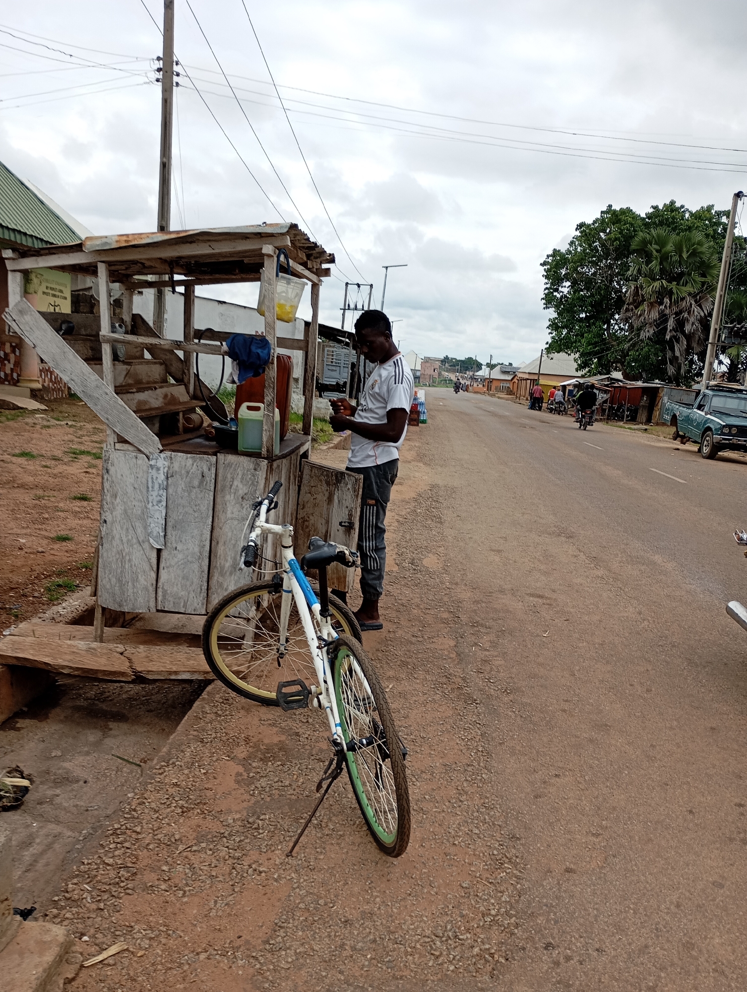 The image shows a man standing next to a makeshift wooden stall on the side of a paved road in Shendam, Nigeria. A bicycle is parked in front of the stall. The man is wearing a white t-shirt with black stripes on the sleeves and dark pants. He is looking down at a mobile phone in his hands. The stall appears to be selling various items, including containers and what looks like a water dispenser. In the background, there are other stalls, some buildings, and vehicles on the road, including a green truck and motorcycles. The sky is overcast. The scene depicts a typical street view with commercial activity on a roadside in Nigeria.