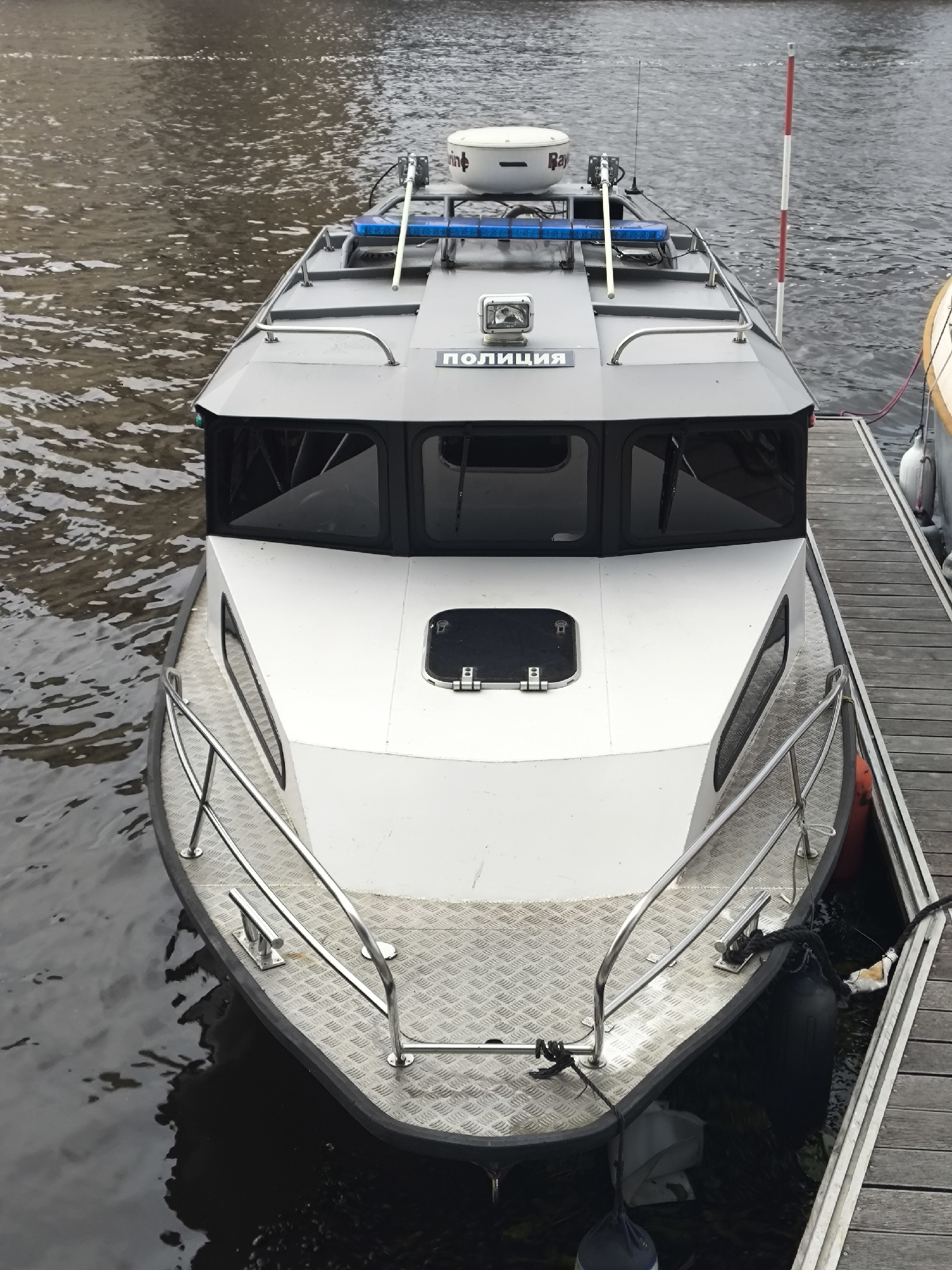 This is a high-angle, full shot of a police patrol boat docked next to a wooden pier. The boat is dark gray and white with black-tinted windows. It has a radar dome and a blue light bar on the roof. The word "ПОЛИЦИЯ" (Police) is printed in white lettering on the side of the cabin. The pier is made of weathered gray wooden planks. The water is dark and choppy. A second boat, partially visible, is docked to the right of the police boat. The scene appears to be in a harbor or marina.