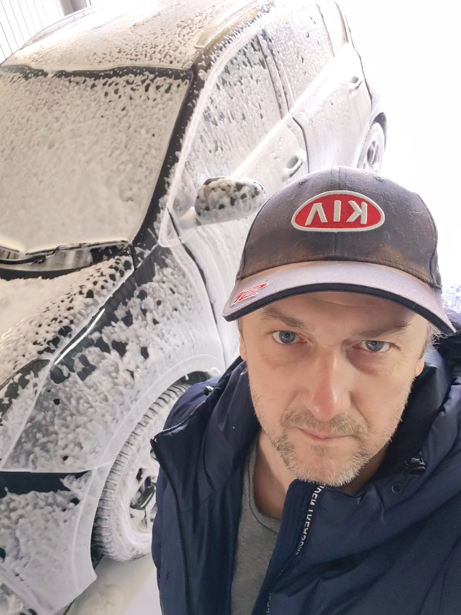 A middle-aged man is taking a selfie in front of a car covered in soap suds, presumably at a car wash. He is wearing a dark blue jacket and a dark baseball cap with a red "KIA" logo on the front. The location is likely in the city of Chaikovsky, Russia, given the logo on the cap. The man's face is clear, showing a stubble beard and light-colored eyes. The car, partially visible behind him, is completely covered in white foam. The background appears to be a bright, possibly enclosed area, consistent with a car wash facility.