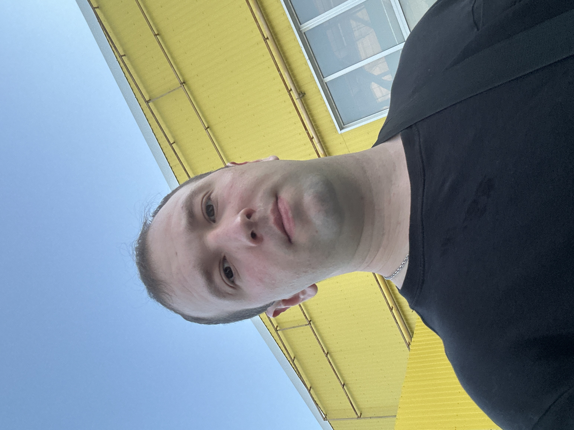 A man with light skin and short hair is positioned looking down at the camera. He's wearing a black t-shirt and a silver chain around his neck. The background is a combination of the sky and a yellow structure, possibly a building. The structure has evenly spaced beams and a window. Given the prompt, the location is speculated to be the city of Chaikovsky, Russia.