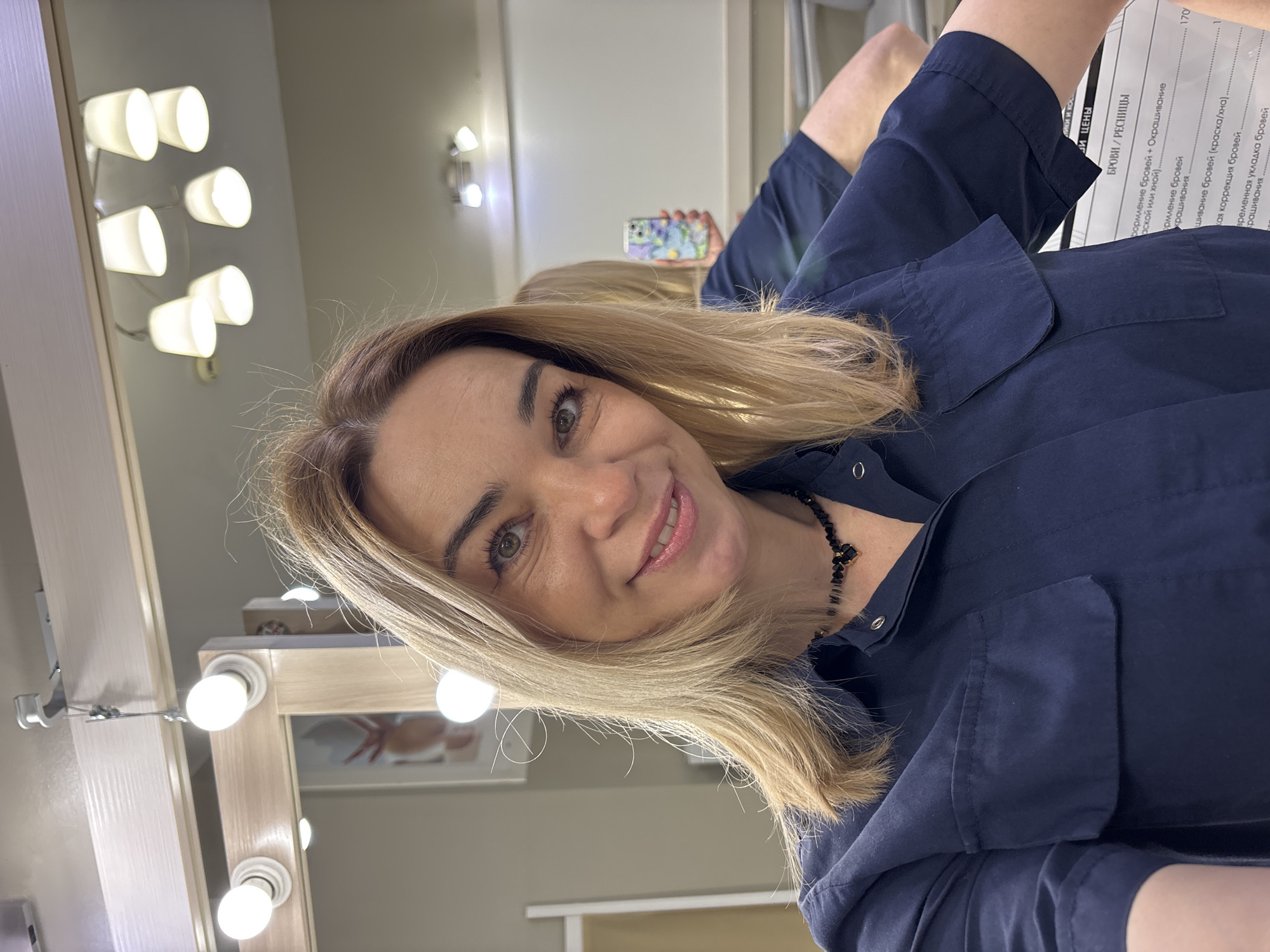 The image is a selfie of a woman with blonde hair, wearing a dark blue button-up shirt and a black necklace with geometric stones. She is smiling at the camera. Behind her, there is a vanity mirror with lights, and a price list on a piece of paper is visible. The text on the list includes words like "БРОВИ / РЕСНИЦЫ" (BROWS/EYELASHES), "Окрашивание" (Coloring), "коррекция бровей" (brow correction).