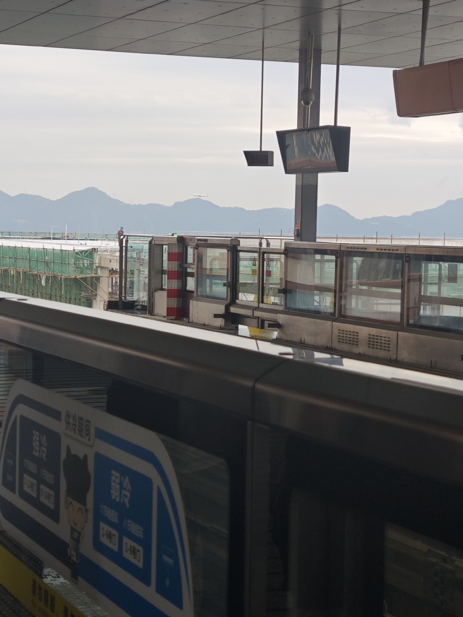 The image shows a train station in Liangjiang, China, with a train at the platform. Construction is underway adjacent to the platform, indicated by scaffolding and a worker in a blue shirt and hard hat standing on the structure. Mountains are visible in the background. Above, there is an overhead structure with hanging monitors. A sign in Chinese on the train car has a cartoon illustration. An airplane can be seen in the sky above.