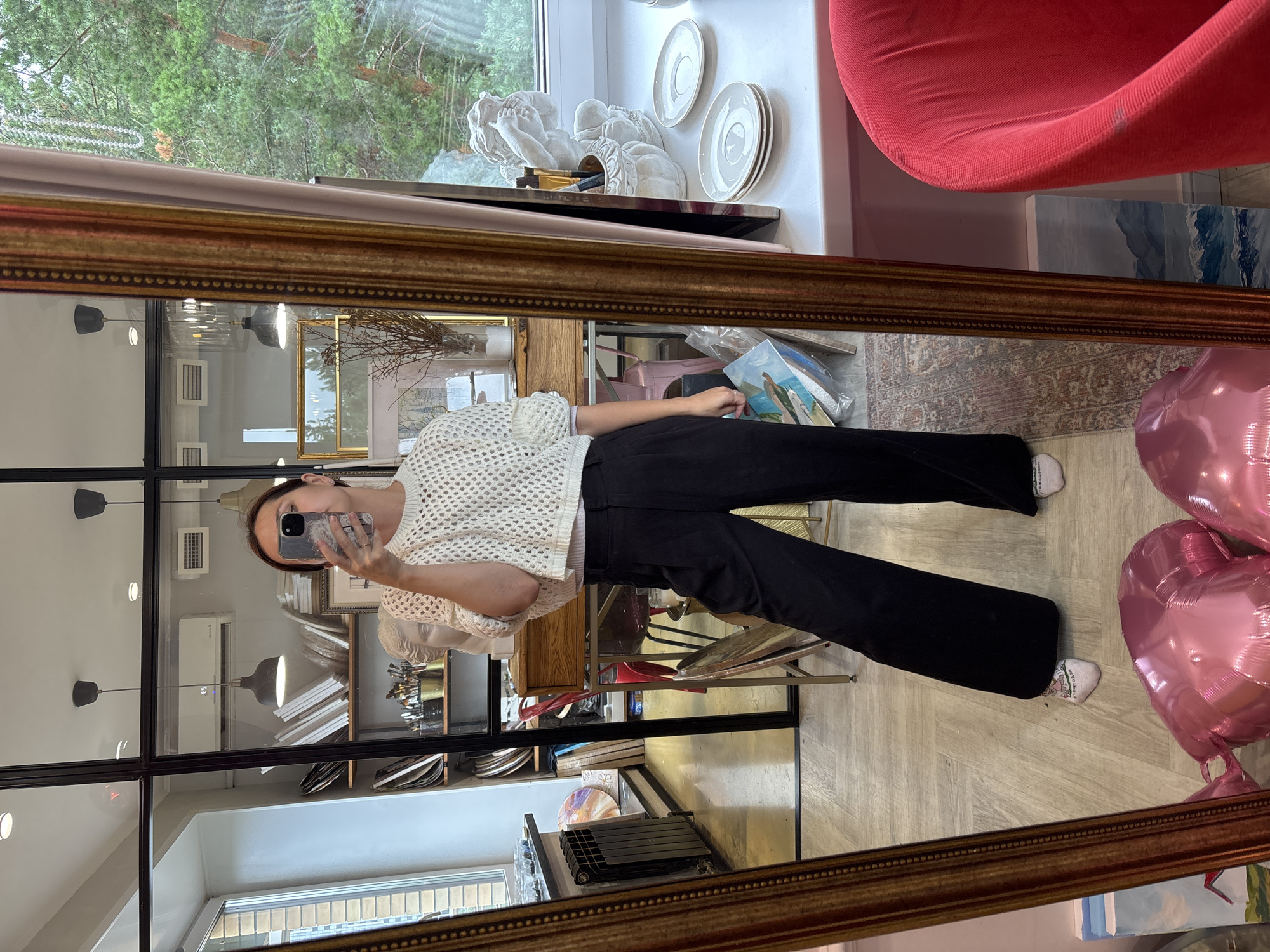 The image shows a woman in a white crocheted top and black wide-leg pants taking a mirror selfie in what appears to be an art studio or shop. She is holding a phone in her right hand. Behind her, shelves are filled with art supplies, canvases, and sculptures. A large ornate mirror dominates the foreground, reflecting the scene. Outside the window, trees are visible, suggesting a location with natural surroundings. The overall environment seems to be a creative space, possibly in Naberezhnye Chelny, Russia, given the context.