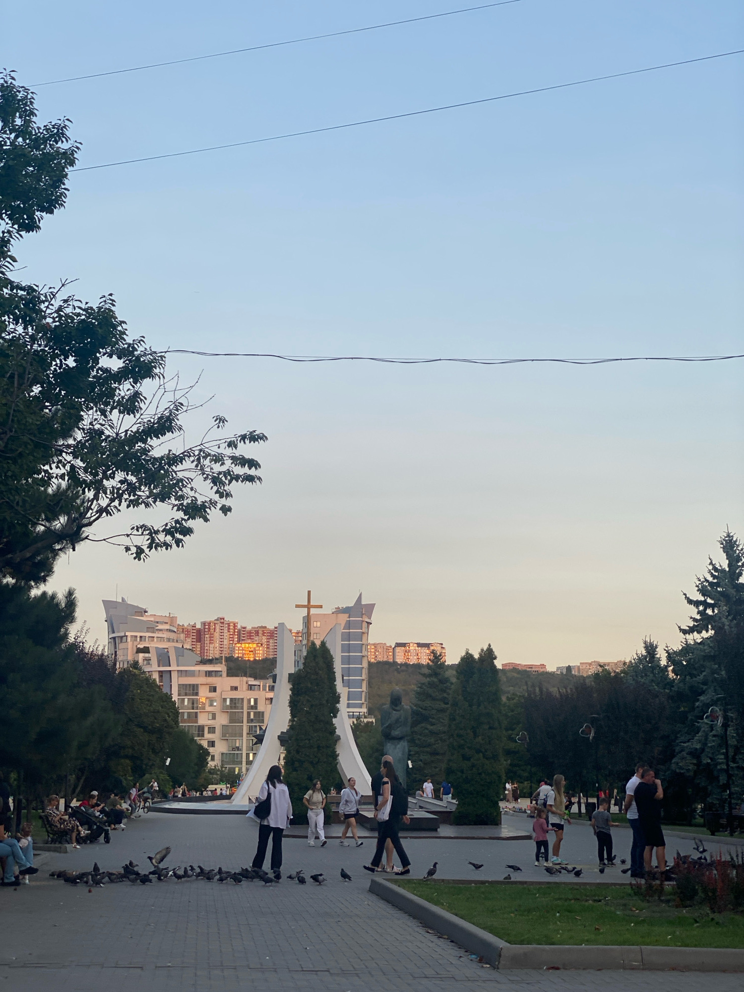 The image depicts a public park scene in Chișinău, Moldova, possibly at the Memorial Complex "Genocide Victims' Memorial" or a similar urban park with a monument and green spaces. In the foreground, a paved pathway is covered with a flock of pigeons, some of which are in motion. Several people are present in the park, with some seated on benches and others walking.
To the left, trees with lush green foliage provide shade. In the mid-ground, a large white monument with a distinctive V-shape is visible, topped with a cross. Adjacent to the monument are several evergreen trees. A statue of a person is also present near the monument. Beyond the park, modern residential buildings and a hillside with more structures are visible in the background. The sky is clear and a pale blue, indicating it is likely late afternoon or early evening. The overall atmosphere appears to be calm and leisurely.