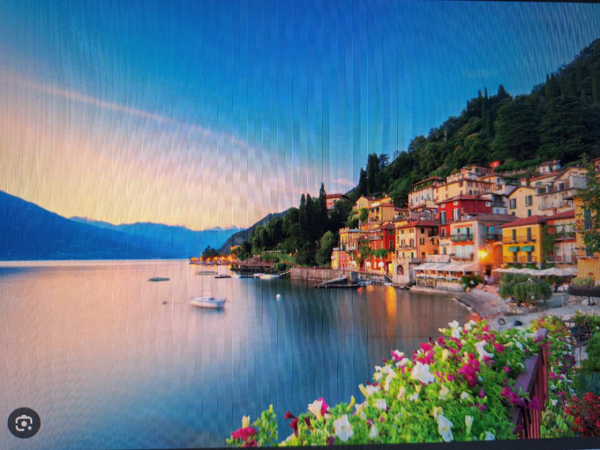 The image depicts a scenic waterfront view of a town nestled on a hillside overlooking a calm body of water, possibly a lake or a bay. The architecture is characterized by colorful, multi-story buildings with red-tiled roofs, suggesting a European setting. These buildings cascade down the hillside, interspersed with lush green trees. In the foreground, a vibrant display of flowers, predominantly white and pink, adds a touch of natural beauty. Several boats are visible on the water, including a white sailboat. The sky transitions from a pale orange and pink near the horizon to a deep blue above, indicating either sunrise or sunset. The scene conveys a peaceful and picturesque atmosphere. This location is not in Varna, Bulgaria.