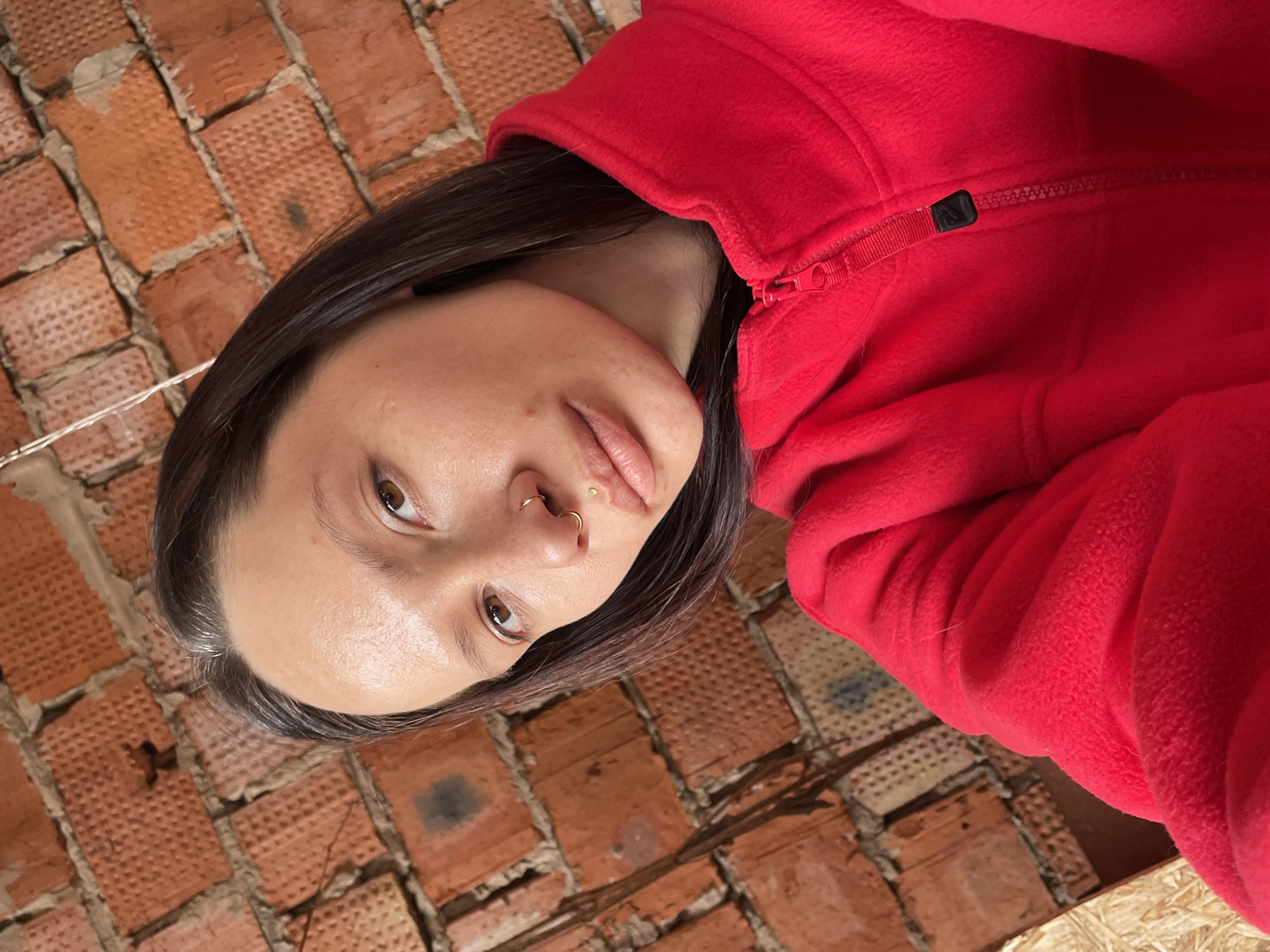 A woman with dark hair, light skin, and a gold nose ring is wearing a red fleece jacket. She is looking slightly upward at the camera. The background consists of an exposed brick wall. The location and city of Oktyabr'skii, Russia, cannot be verified based on the image alone.