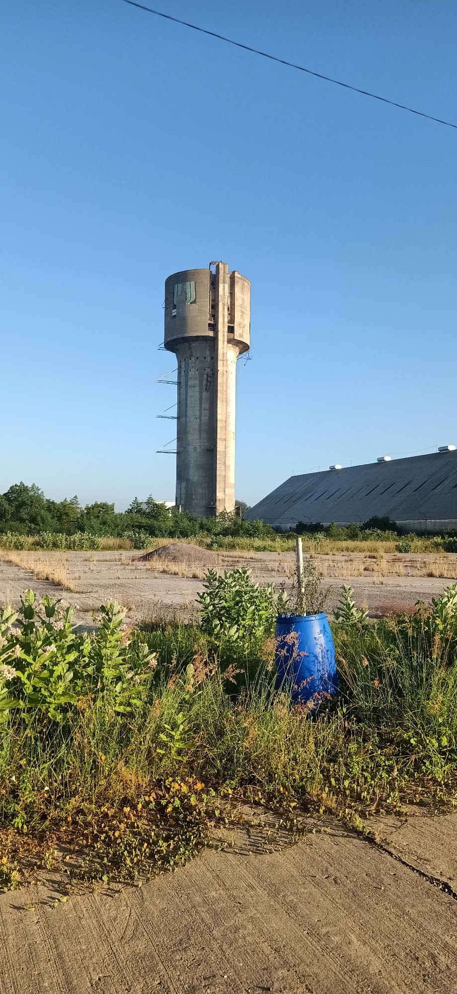 In Keudekruenggeukueh, Indonesia, a tall, cylindrical concrete water tower stands against a clear blue sky. The tower has a weathered appearance with visible horizontal lines and some scaffolding-like structures attached to its side. At its base, a blue plastic barrel is situated amidst overgrown green vegetation and dry grasses. In the background, a large, angular, grey-roofed building with visible beams and supports is visible. The ground in the foreground is a rough, grey concrete surface, with a patch of dry, cracked earth and scattered dry vegetation behind the immediate green growth.