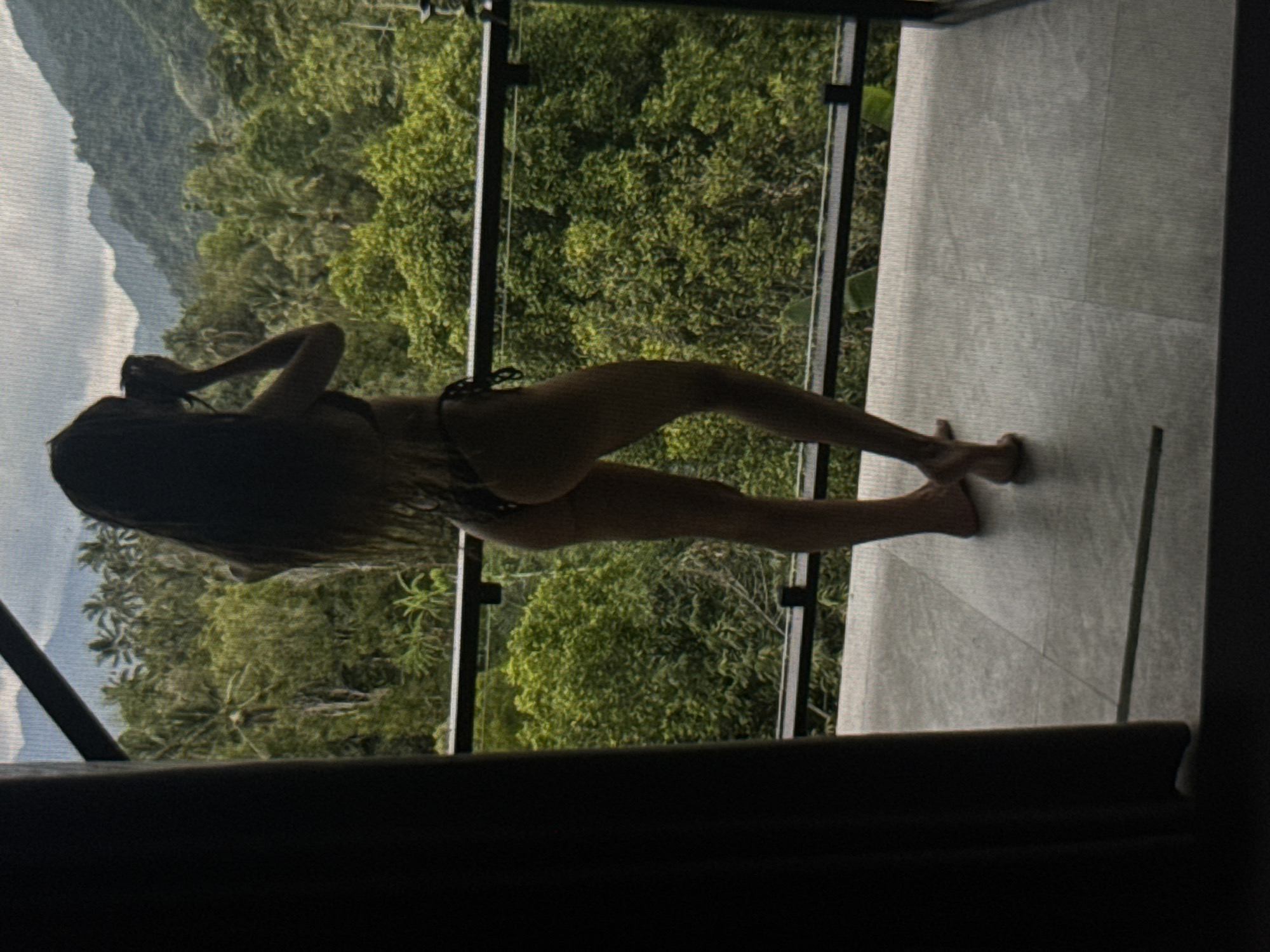 A person is standing on a balcony, facing away from the camera, holding a camera to their face as if taking a picture. They are wearing a bikini. The balcony overlooks a dense jungle landscape with mountains in the distance. The area appears to be Kuta Selatan, Indonesia, known for its lush scenery and tropical climate. The lighting suggests it is daytime, with soft natural light illuminating the scene.