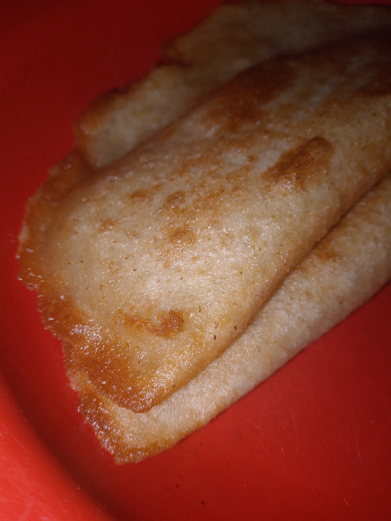 The image shows two crepes stacked on a red surface. The crepes have a light brown color with a slightly golden hue around the edges. The surface texture of the crepes appears slightly porous. No people, specific location in Juan Griego, Venezuela, or other objects are visible. The action is that of placing the crepes on the red surface. No further details can be determined from the image.