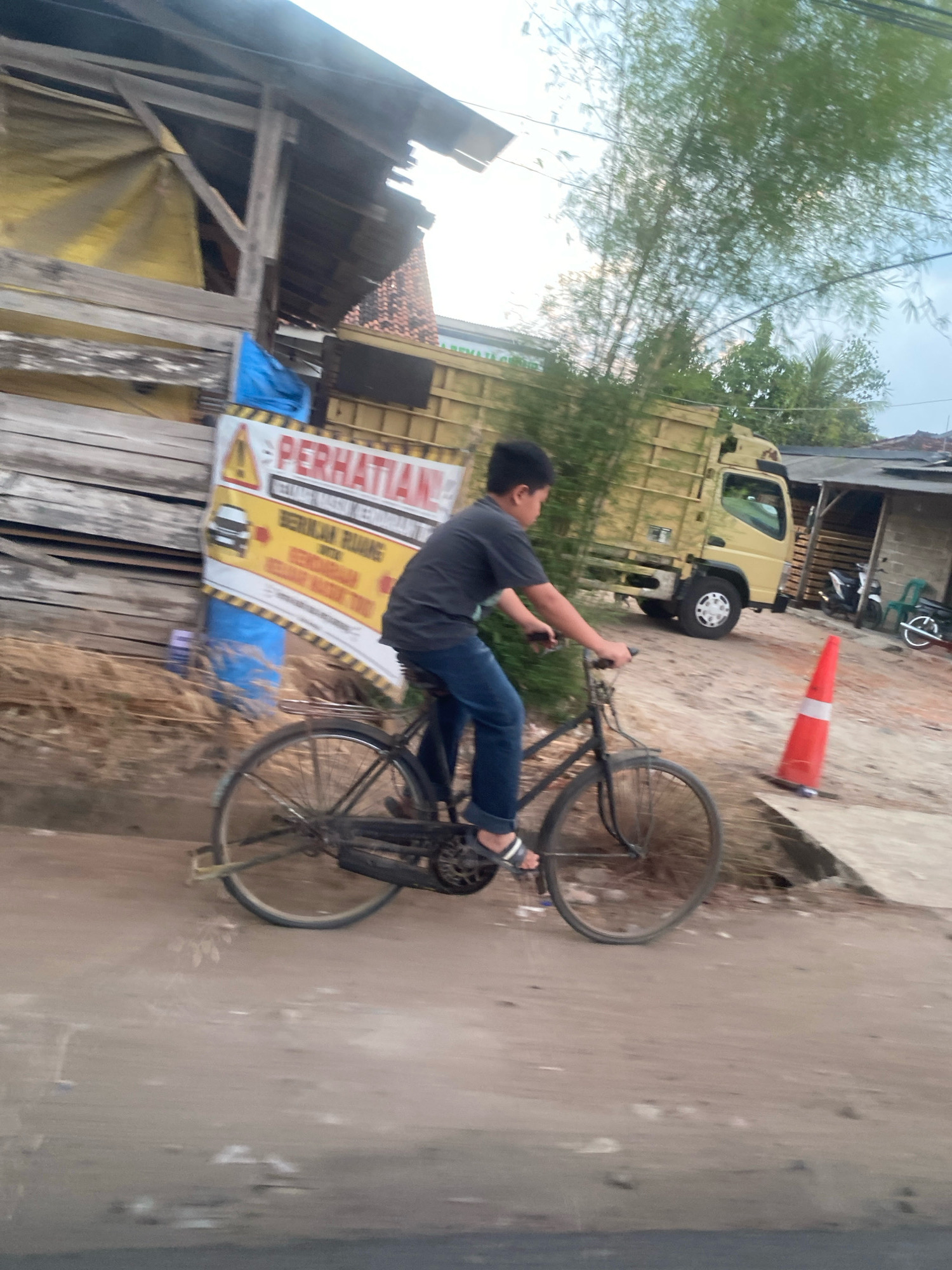 In Banjar Sari, Indonesia, a young boy is riding a bicycle on a dirt road. He is wearing a dark t-shirt and blue jeans. The bicycle is black and has a metal frame. In the background, there is a yellow truck and a building with a corrugated metal roof. There is also a sign with the word "PERHATIAN" which means "ATTENTION" in Indonesian. The area appears to be under construction or in a state of disrepair, with visible building materials and debris. The lighting suggests it is either early morning or late afternoon.