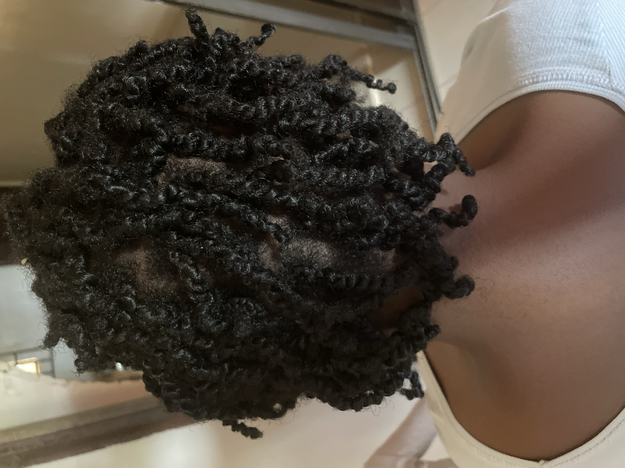 This is a close-up, back-view, medium shot photograph of a person's head and shoulders. The person has dark, curly hair styled in twists, which are covering most of their head. The hair appears to be dark brown or black. The person is wearing a white top with a textured neckline. The background is blurred but suggests an indoor setting with neutral-colored walls and possibly a window or reflective surface on the right side. There is no identifiable indication of the city of Kakau, Nigeria, or any specific urban environment. The image focuses entirely on the person's hair and upper body.