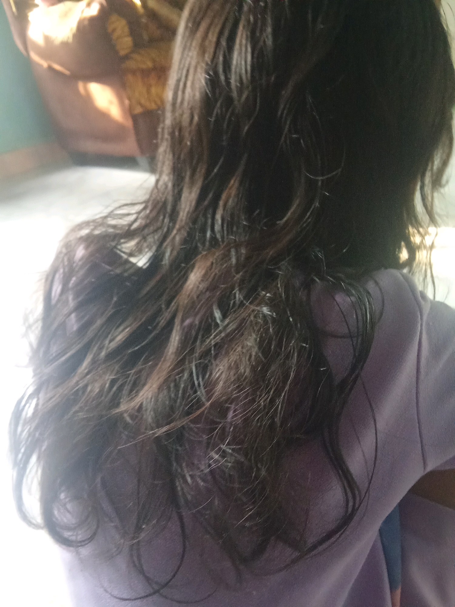 The image, taken in Cadasari, Indonesia, is a close-up, rear-facing shot of a person, likely a child, with dark, wavy hair. The subject is wearing a light purple garment, possibly a t-shirt or sweatshirt. The lighting suggests the photo was taken indoors near a window, with a bright light source creating a strong glare on the left side of the image and illuminating the background which includes a hint of a patterned fabric. The focus is primarily on the subject's hair and upper back.