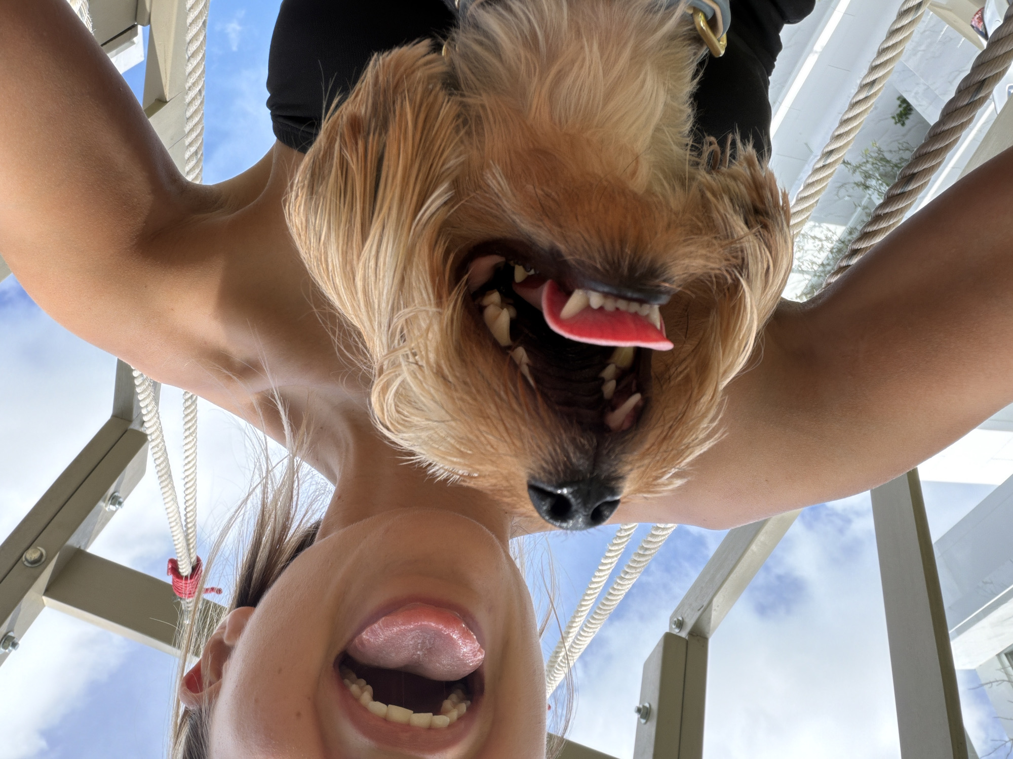A close-up, selfie-style photograph captures a young woman and a small, tan-colored dog positioned in the foreground, against a backdrop of a cloudy sky and a wooden structure. The woman is on the right side of the frame, with her mouth open and tongue sticking out. Her left arm is visible, extended upwards and bent at the elbow. The dog is on the left, directly in front of the woman, also with its mouth open, revealing its teeth and tongue. The wooden structure appears to be part of an outdoor play area, possibly a playground or a hammock frame, with thick ropes attached. The scene is shot from a low angle, looking upwards. The location is identified as Thep Krasatti, Thailand.