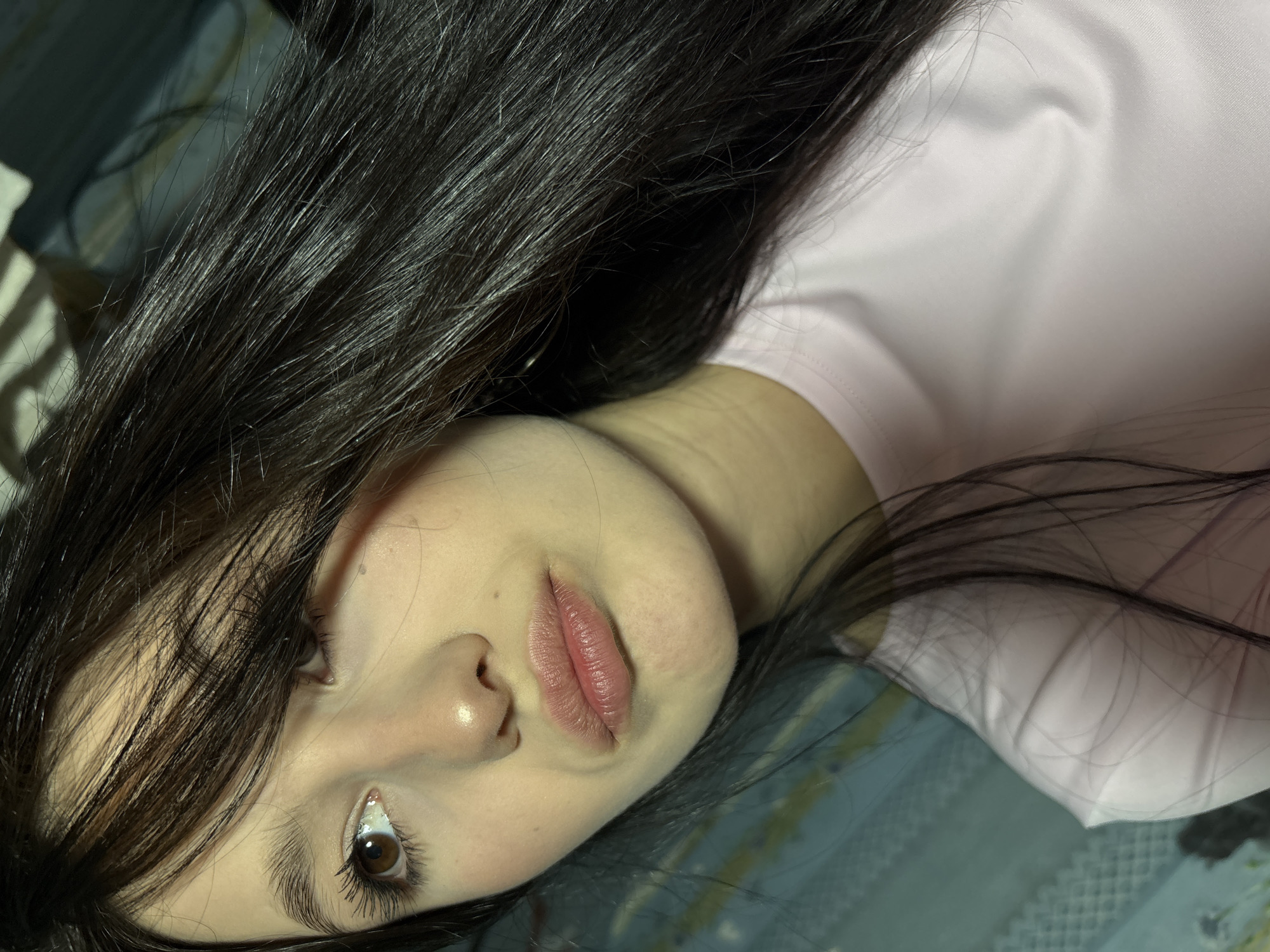 A close-up, chest-up portrait features a young woman with long, dark brown hair, parted on the side, with strands partially obscuring the right side of her face. She has brown eyes, with visible eyelashes, and a light complexion. Her lips are closed and slightly pursed, indicating a pouting expression. She is wearing a light pink, possibly satin or silky, crew-neck top. Her gaze is directed towards her left, off-camera. The background is out of focus, revealing an indoor environment with a light blue, vertically striped or patterned surface on the left and darker, indistinct shapes to the upper right. There are no visual cues within the image to confirm the location as the city of Фоки, Russia.