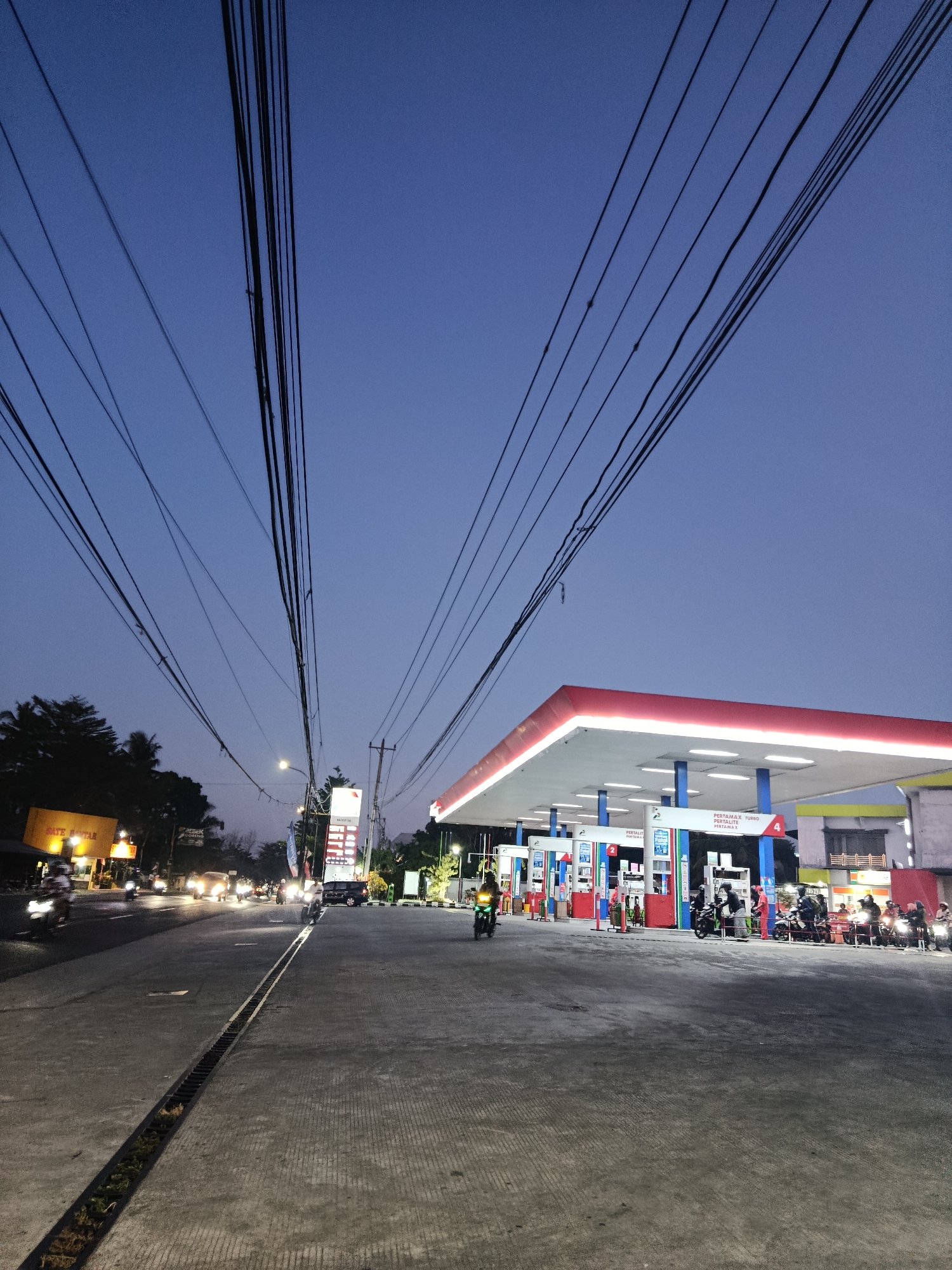 This is a vertical, full-shot photograph taken at dusk on a street in Sedayu, Indonesia. The image is dominated by a clear, twilight blue sky and a network of black power lines stretching across the upper portion of the frame.
In the foreground, a paved road and sidewalk are visible. A metal grate runs along the edge of the sidewalk, with some vegetation visible in the trench beneath it.
To the right of the road is a brightly lit gas station with a red and white canopy. Multiple fuel pumps are arranged under the canopy, some with people on motorcycles in front of them. A tall, illuminated signpost stands near the gas station, displaying prices and fuel types.
On the left side of the road, a row of buildings with illuminated signs is visible. One sign, in yellow, reads "SATE KALTAR" in red lettering. Further down the road, headlights from several vehicles, mostly motorcycles, illuminate the scene.
People are present at the gas station, primarily on motorcycles. Some are interacting with the pumps, while others appear to be waiting. A few individuals are also visible on the sidewalk.
The overall impression is of a busy street scene in a semi-urban area at the end of the day, with artificial lights beginning to illuminate the surroundings. The presence of the gas station and the numerous motorcycles suggests this is a common thoroughfare.