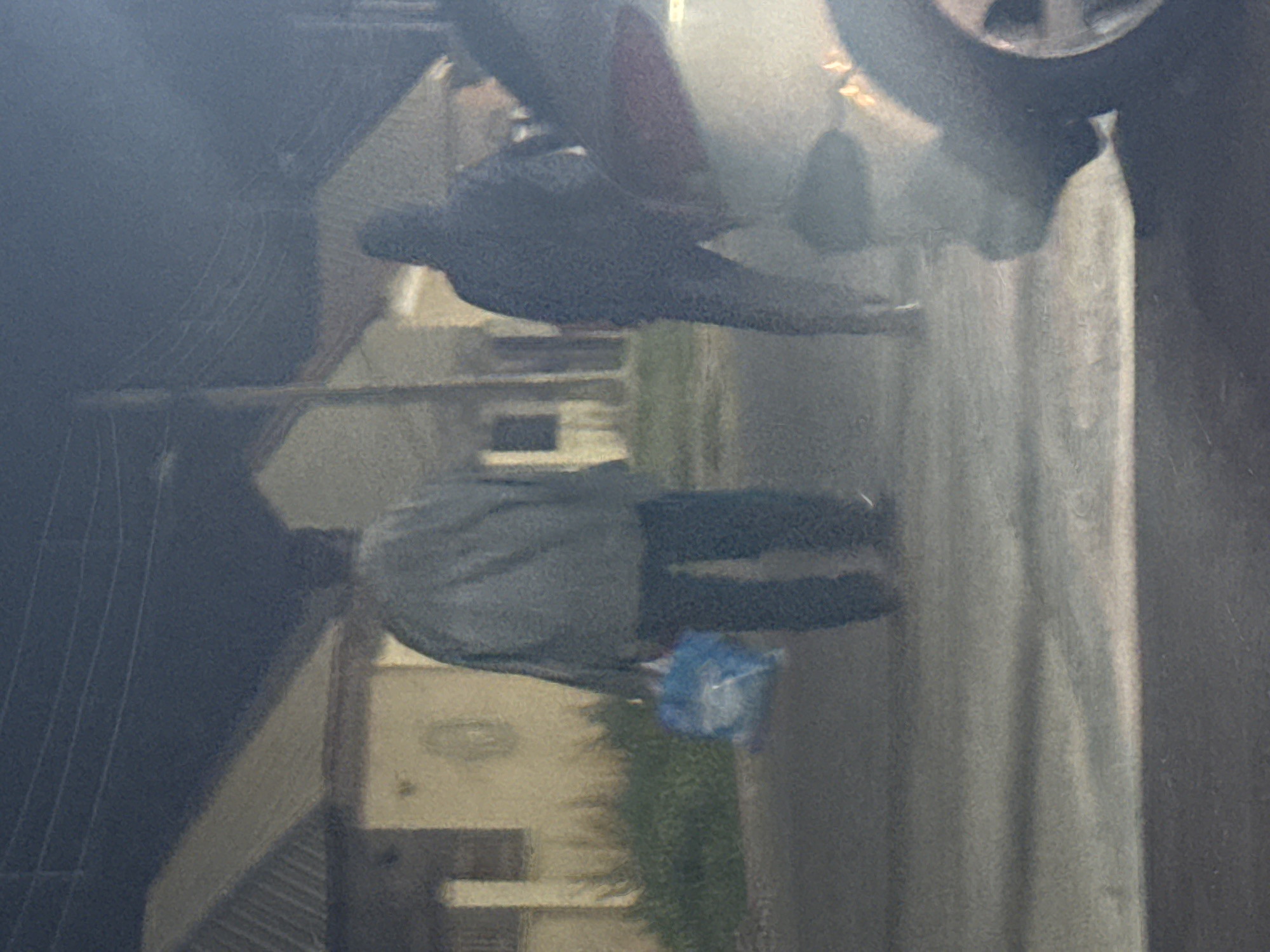Two individuals are walking away from the camera on a street in Abuja, Nigeria. The person on the left is wearing a grey jacket and dark pants, and is carrying a blue plastic bag in their left hand. The person on the right is wearing a dark hooded jacket. Buildings with peaked roofs are visible in the background, along with some vegetation. A vehicle is partially visible in the foreground on the right side of the image. The scene appears to be illuminated by streetlights or ambient light, creating a somewhat grainy and dark atmosphere.