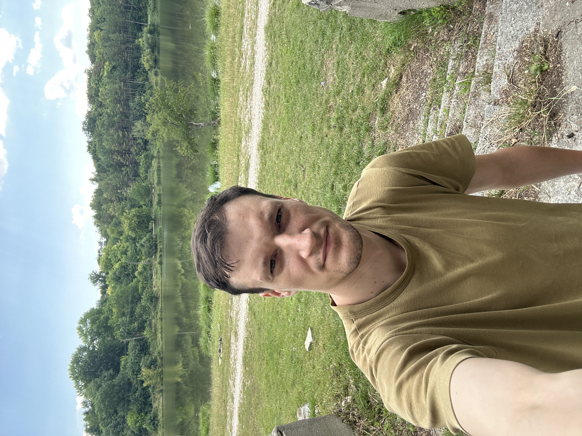 In Makiv, Ukraine, a man takes a selfie outdoors by a lake. He is wearing a light brown t-shirt and stands in the foreground on a stone staircase, looking directly at the camera. His arm is extended, holding the phone. Behind him, a calm lake reflects the lush green trees and blue sky. The environment is natural, with grass, gravel paths, and dense woodland surrounding the water.