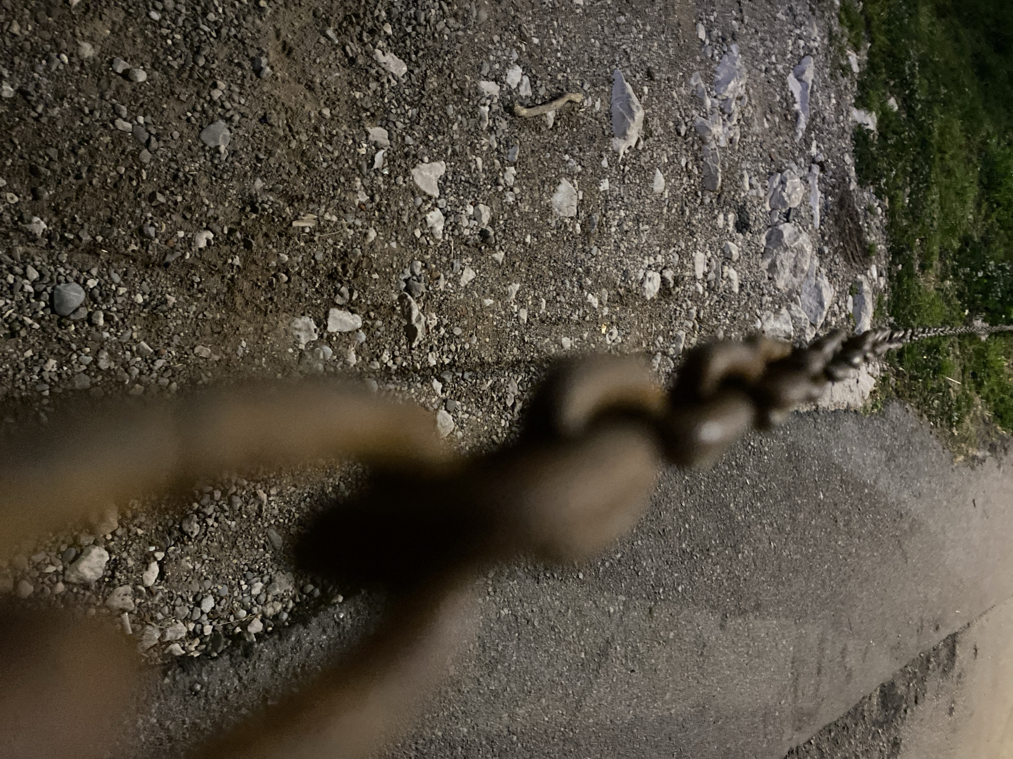 The image shows a ground-level view featuring a combination of rough gravel and asphalt. Patches of green grass are visible at the top-right corner. A metal chain, partially in focus, extends diagonally across the frame. The setting appears to be outdoors, possibly in Budva, Montenegro. The chain could be part of a barrier or support structure. No people or animals are visible. The scene is dimly lit.