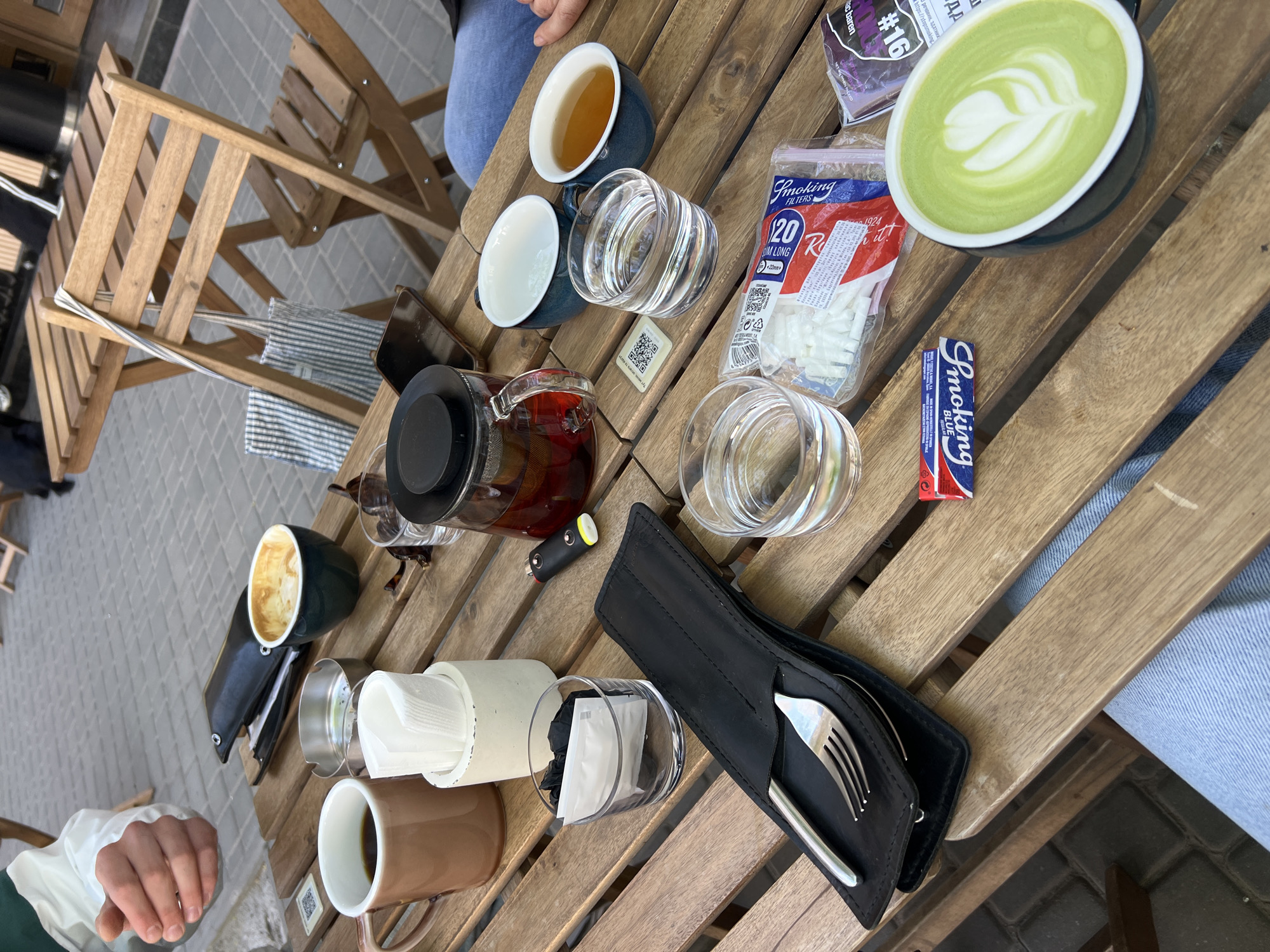 This image shows a tabletop scene in Odesa, Ukraine, on a wooden table with chairs, indicating a cafe or outdoor seating area. Several objects are arranged on the table. There are multiple cups and glasses of varying shapes and sizes, some filled with liquid that appears to be tea and another with a green drink adorned with latte art. A glass teapot with tea is also present. A package of cigarette filters, a box of rolling papers, a cutlery holder with a fork and spoon, and a cylinder filled with tissues are also on the table. The scene also shows parts of two people, one in jeans and another wearing a white shirt and dark green sleeve. The presence of smoking-related items suggests that people are relaxing or socializing over drinks and possibly cigarettes. The setting is a casual outdoor location, with some chairs and stone flooring visible around the wooden furniture.