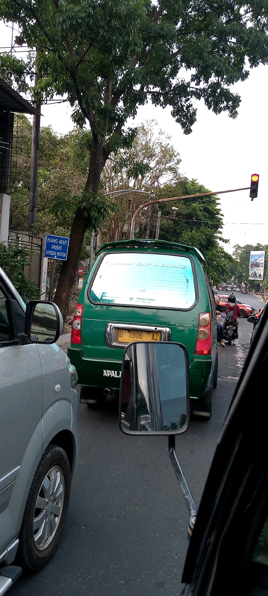 The image shows a view from inside a vehicle, looking out at traffic on a street. The main focus is a green car with a license plate that reads "D 1906 AZ". There is also a sign on the left that says "RUANG HENTI KHUSUS SEPEDA MOTOR," which translates to "SPECIAL WAITING AREA FOR MOTORCYCLES." The scene appears to be in a city, with trees and buildings visible in the background. A traffic light is visible in the upper right of the image, showing a yellow light. Other vehicles and motorcycles are also present in the traffic. The overall impression is of a typical urban traffic scene in Indonesia.