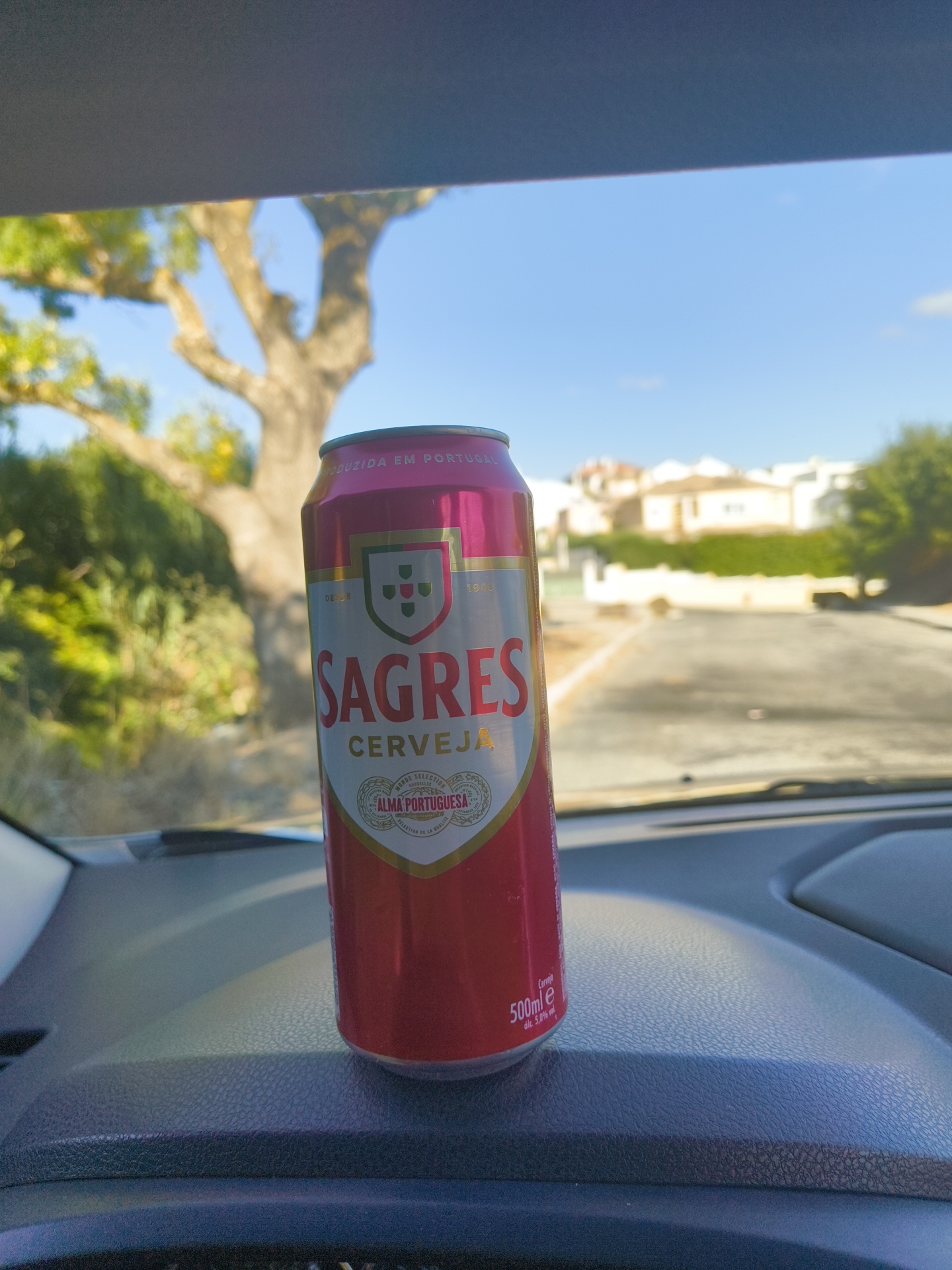 A red and silver can of Sagres Cerveja sits on a car's dashboard in a sunlit outdoor environment. The can is the primary subject, with its label clearly visible. In the background, a tree with bare branches and green foliage is visible, along with a street and houses, suggesting a suburban or residential area. The sky is blue and clear. The scene is likely in Cascais, Portugal, given the brand of beer and the visual cues of the environment. No people are present in the image.