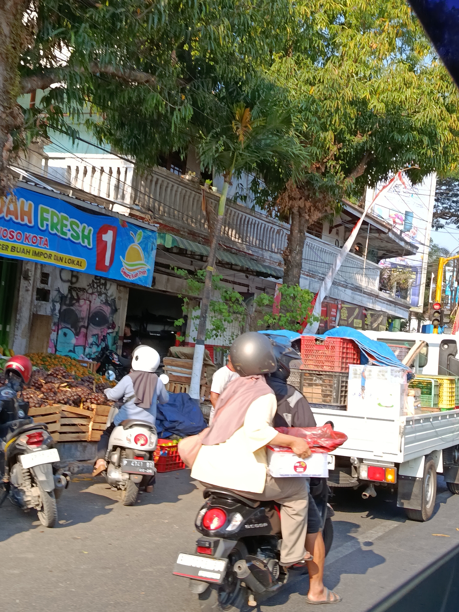 In the city of Bondowoso, Indonesia, on a street with fruit stalls, two people ride on a motorcycle. The driver wears a dark helmet, and the passenger, in a tan shirt and brown pants, holds a white box and a red bag. The motorcycle is behind a white flatbed truck carrying red and blue crates. In the background, a fruit stall displays various fruits in wooden crates. Another motorcycle, with a red helmet on its handlebars, is parked nearby. The street is paved, and trees line the side of the road. The scene is captured in daylight.