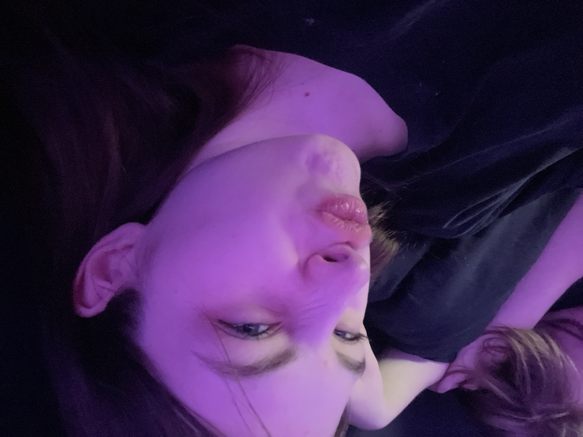 A photograph depicts a woman’s face and upper body, rotated 90 degrees counter-clockwise. Her face is illuminated with a pinkish-purple hue, likely from artificial lighting. She has fair skin, dark hair, and is wearing a dark-colored garment. The background is indistinct and dark. The image provides no indication of being taken in St. Petersburg, Russia.