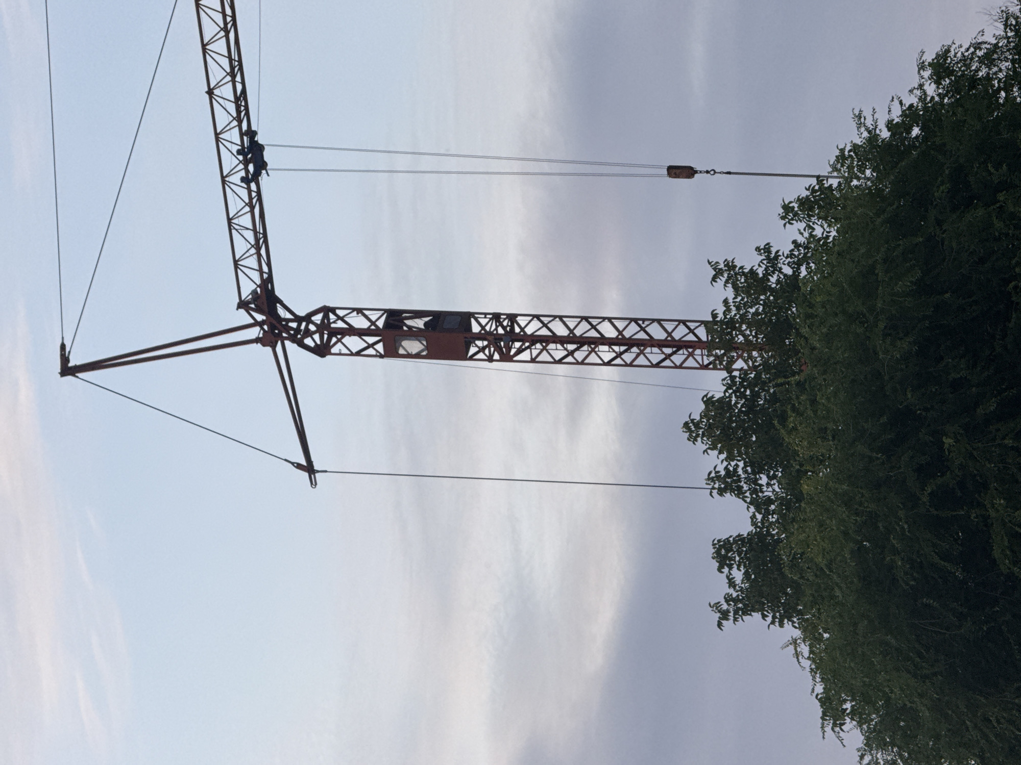 A tall, red, lattice tower crane is positioned behind a dense canopy of green trees. The crane has a horizontal boom extending to the right. A small, enclosed operator's cab is visible on the vertical section of the crane. Two cables hang from the horizontal boom, one with a cylindrical weight attached. The sky behind the crane is a pale blue with wispy, white clouds. The image is taken from a low angle looking up. The location is Ialoveni, Moldova.