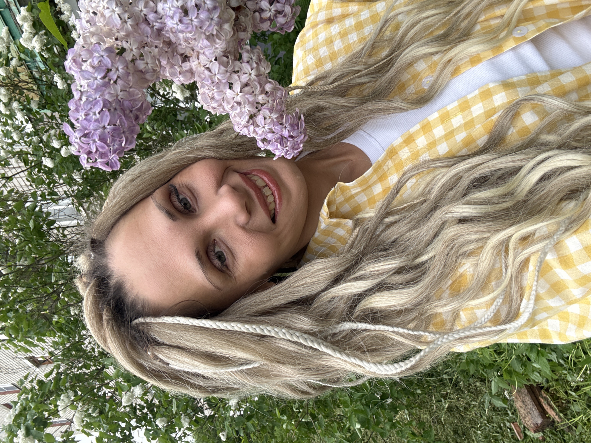 A woman with light blonde hair is smiling at the camera. She is wearing a white t-shirt under a yellow and white checkered button-down shirt. There are lilac flowers above her head, and greenery is visible in the background. Part of a building can be seen in the distance. The scene is in the city of Rusanovo, Russia.