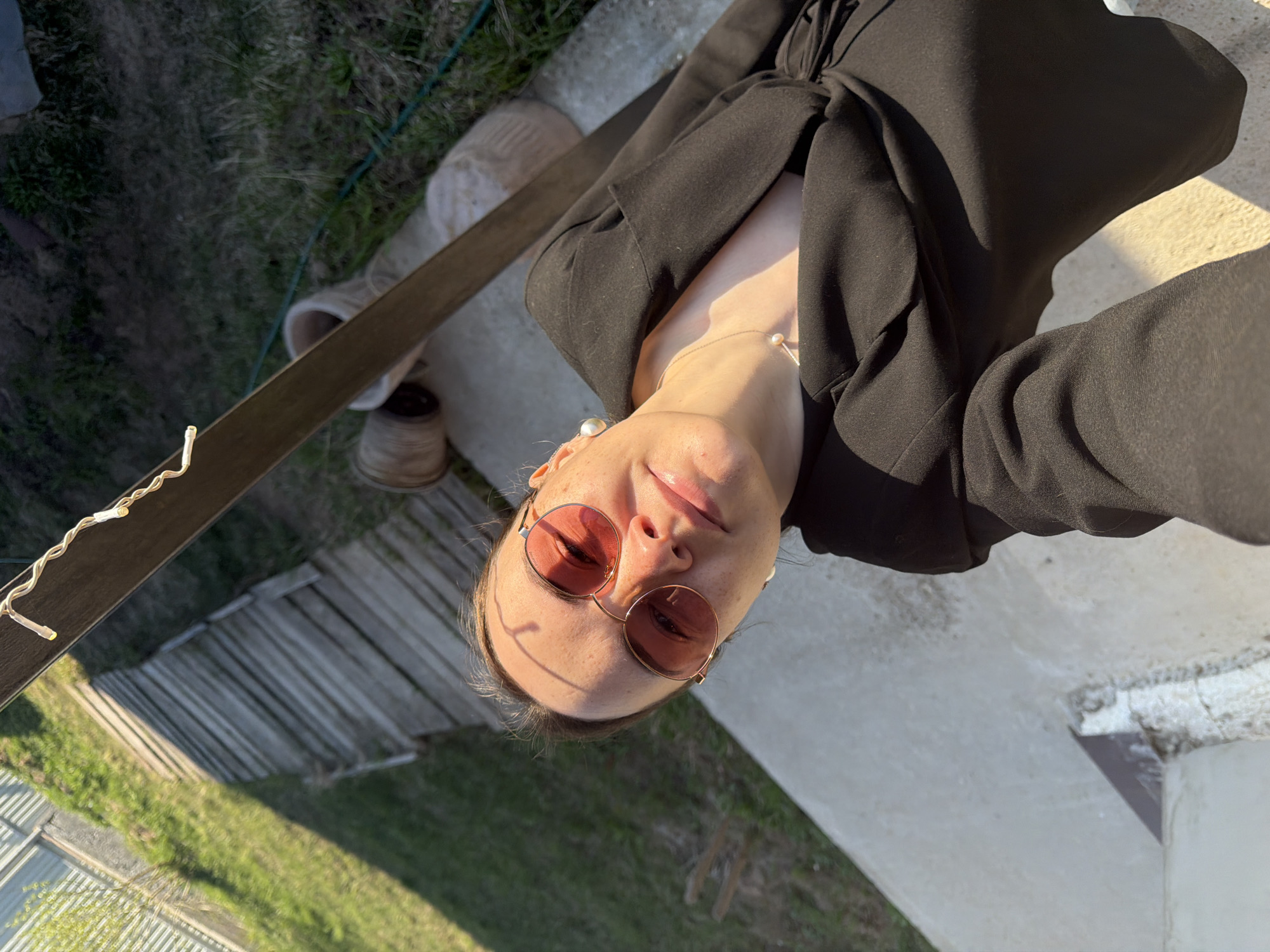 A person wearing round, reddish-tinted sunglasses and a black blazer is the central subject of this eye-level, rotated outdoor selfie. The individual is also wearing a pearl necklace and has earrings that match.
The setting appears to be a yard or garden, as visible in the background are sections of a wooden fence, green grass, and a hose. A wooden plank with string lights attached is on the left side of the frame. A concrete structure is in the background as well.
It is impossible to confirm the city of Chaikovsky, Russia, based on the visual information.