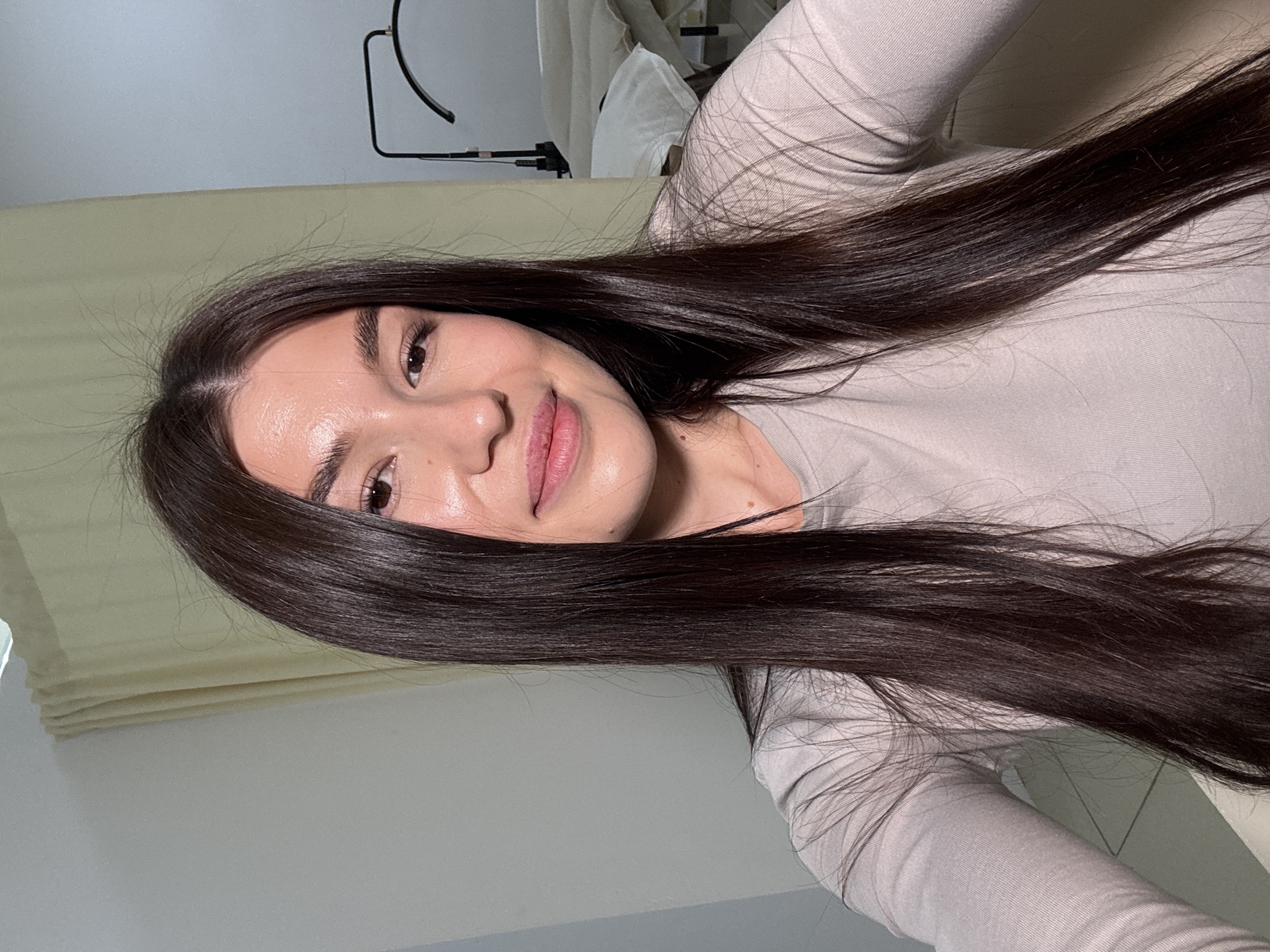A young woman with long, dark, straight hair, parted on her left side, is the central subject of this close-up selfie. She is looking directly at the camera with a soft, gentle smile, her lips slightly parted to reveal a hint of pink. Her complexion is fair and appears luminous. Her eyes are dark, and she has visible eyelashes, suggesting light makeup. Her dark eyebrows are well-defined.
She is wearing a plain, long-sleeved top in a light beige or greyish hue, possibly a ribbed or knitted fabric. Her hair cascades over her shoulders and upper torso.
The setting appears to be indoors. In the background, to her left, there's a light-colored wall and what looks like a light-colored curtain or blind. To her right and behind her, a light-colored surface, possibly a bed or sofa with light bedding/upholstery, is visible. Above her head, a dark, curved object, resembling part of a lamp stand or ring light, is present against a lighter wall or ceiling. The overall lighting is bright and even, consistent with indoor lighting conditions. There is no visible text in the image. The image captures a serene and pleasant moment.