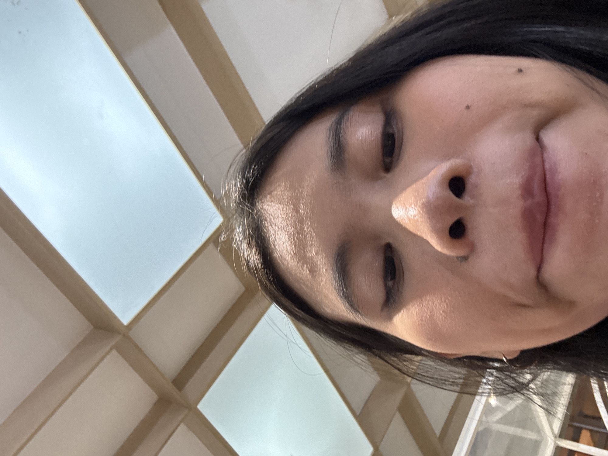 This is a close-up, low-angle selfie of a person's face, taken outdoors. The person has dark hair, and their face is partially obscured by the angle of the shot and their hair. The background features a geometric pattern of light and tan-colored panels, possibly part of an outdoor structure or ceiling, suggesting it might be daytime. The lighting indicates a bright environment, possibly sunny. The person's expression is neutral. The location context provided is Central Jakarta, Indonesia. There is no visible text in the image.