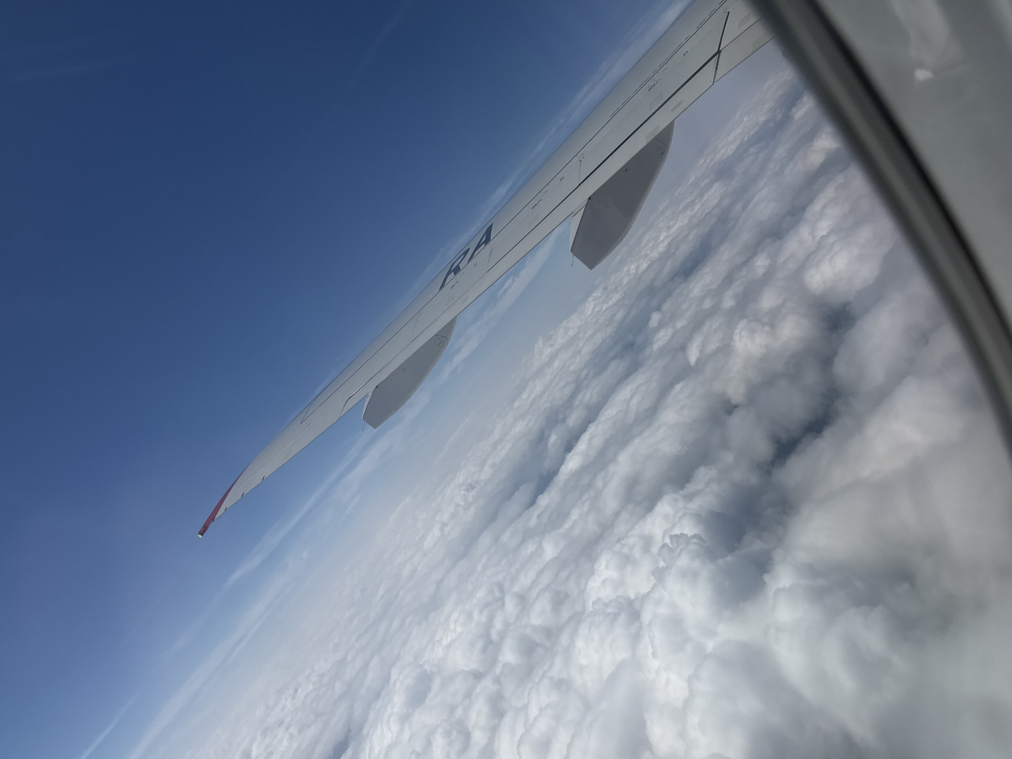 This is an aerial view from an airplane window looking out over a thick layer of white, fluffy clouds under a clear blue sky. The wing of the airplane is prominently visible on the right side of the frame, extending diagonally across the image. The wing is white with a red tip and has some dark lettering, partially obscured, that appears to read "RA". The image does not contain any information about the city of Staroe Mart'yanovo, Russia.