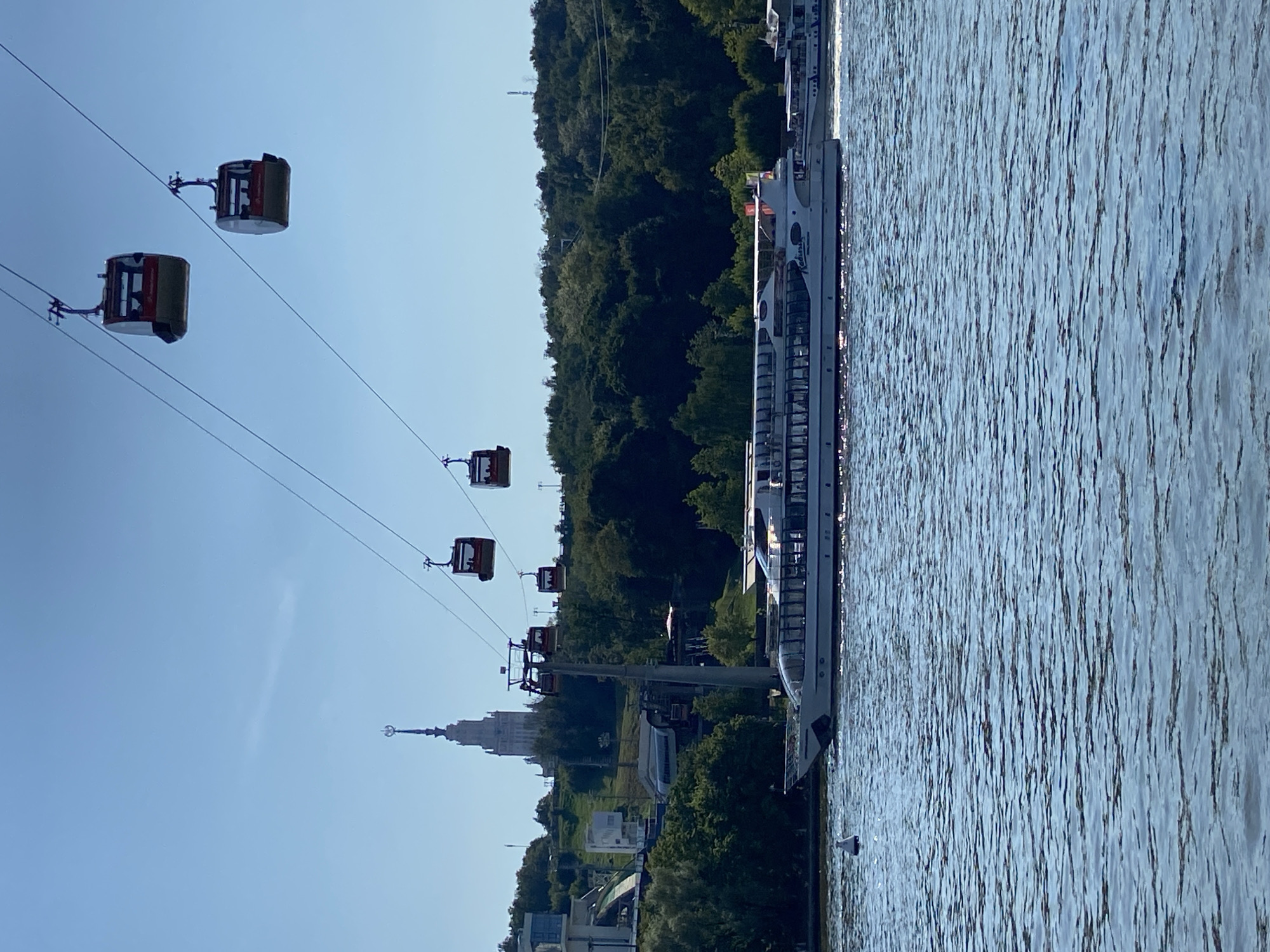 A clear, sunny day is depicted in Moscow, Russia, featuring a wide river with a large white passenger boat cruising on it. The boat has multiple decks and appears to be carrying passengers. In the background, a lush green hillside is covered with trees, and a tall, ornate white building with a spire and a star on top is visible at the crest of the hill, characteristic of Stalinist architecture. Above the river and hillside, several red and white cable car cabins are suspended from wires, traversing the sky. The water is choppy, reflecting the sunlight. A white buoy floats in the foreground of the river.