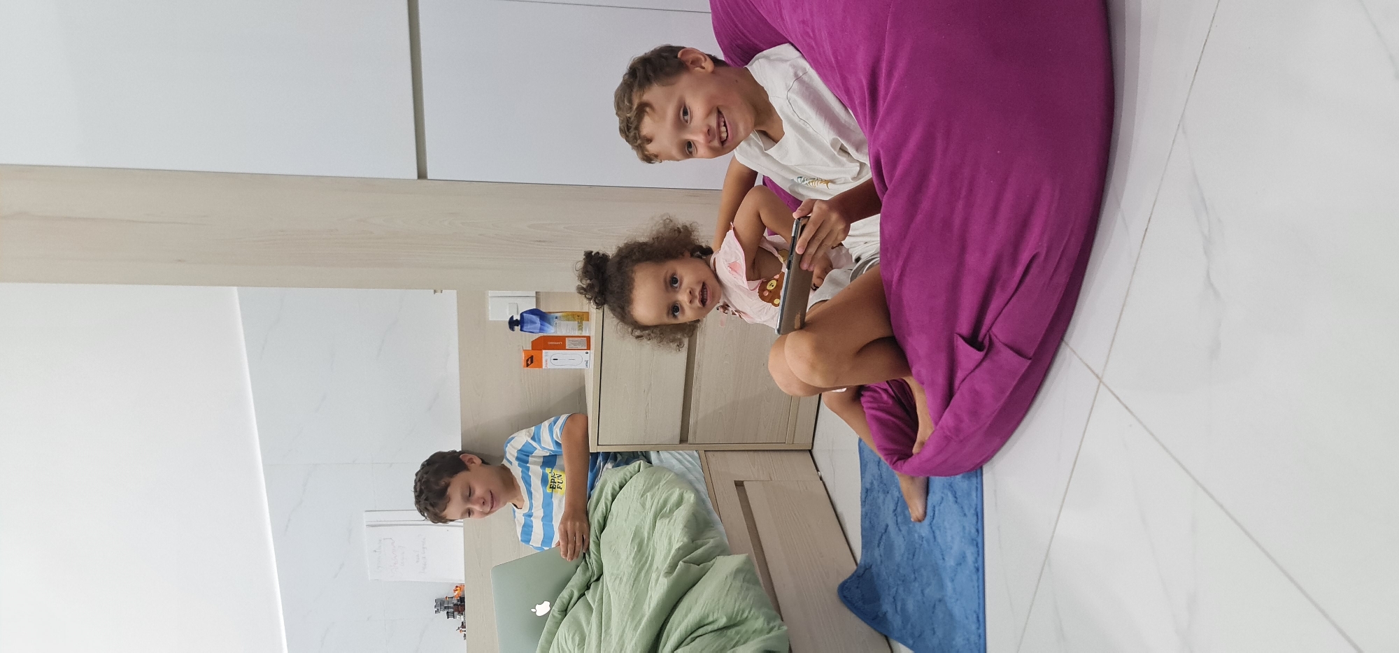 A brightly lit interior space with white and light wood-toned walls and tiled floors contains three children and various objects. The children include one boy working on a laptop in the lower left, a second boy seated with a small girl in a large purple beanbag chair. The girl is holding a cell phone. A blue rug lies near the beanbag chair. To the left of the children in the beanbag is a light wood-toned dresser. The location is possibly in the city of Da Nang, Vietnam.