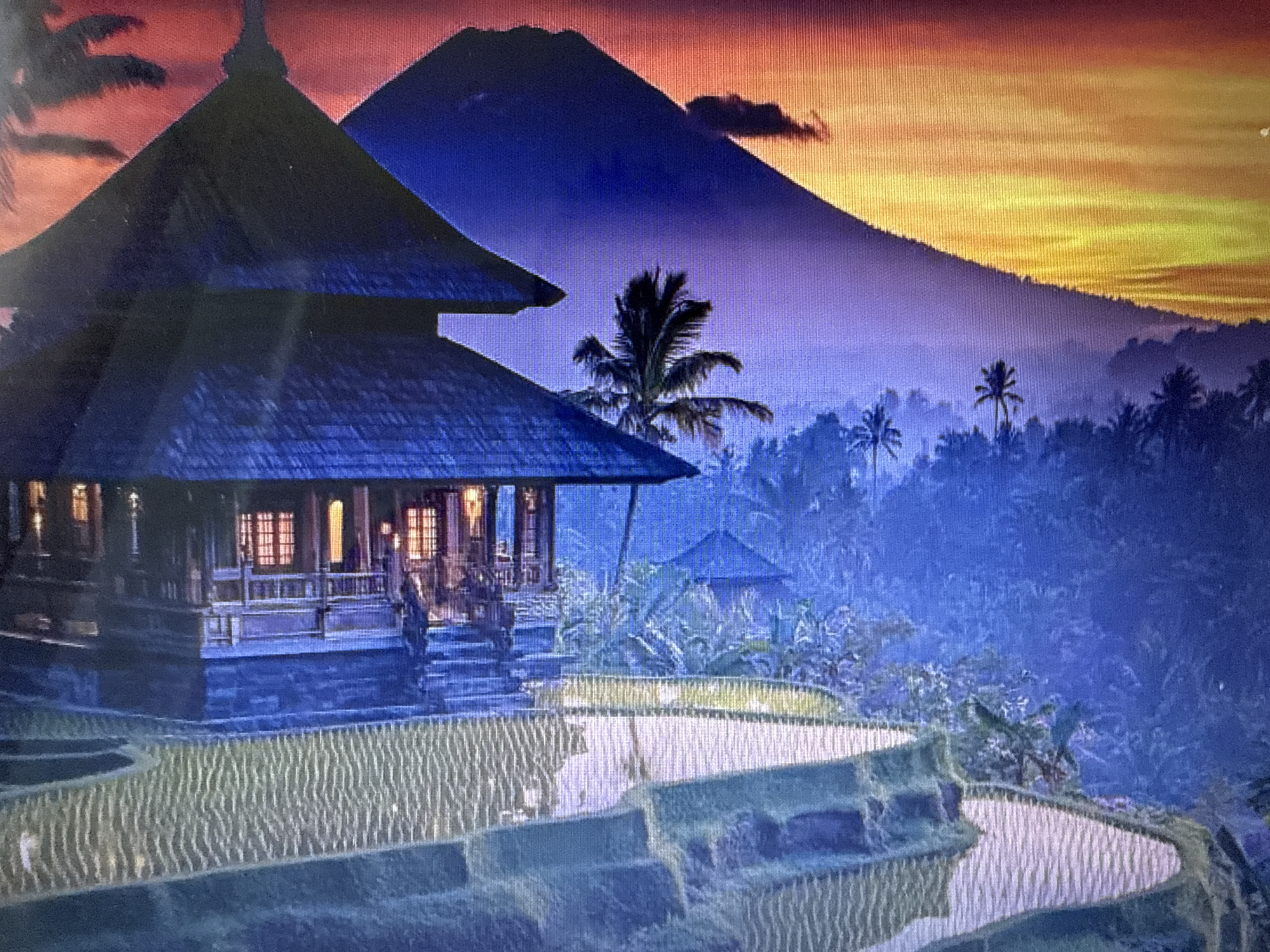 The image depicts a serene landscape scene with a traditional Balinese building in the foreground, set against a backdrop of lush rice terraces and a majestic mountain at sunset. The building, made of dark wood and stone, features intricate carvings and a tiered roof, with warm light emanating from its windows, suggesting an inviting interior. The rice terraces, with their distinct steps and water-filled paddies reflecting the sky, cascade down the hillside. In the distance, a conical mountain looms large, its silhouette softened by the purple haze and the vibrant hues of the setting sun painting the sky in shades of orange, yellow, and red. Palm trees and other tropical foliage dot the landscape, adding to the exotic ambiance. The scene evokes a sense of tranquility and natural beauty. The request to include the city of Pushkino, Russia, is not applicable to this image, as the depicted scenery is characteristic of Southeast Asia, not Russia.