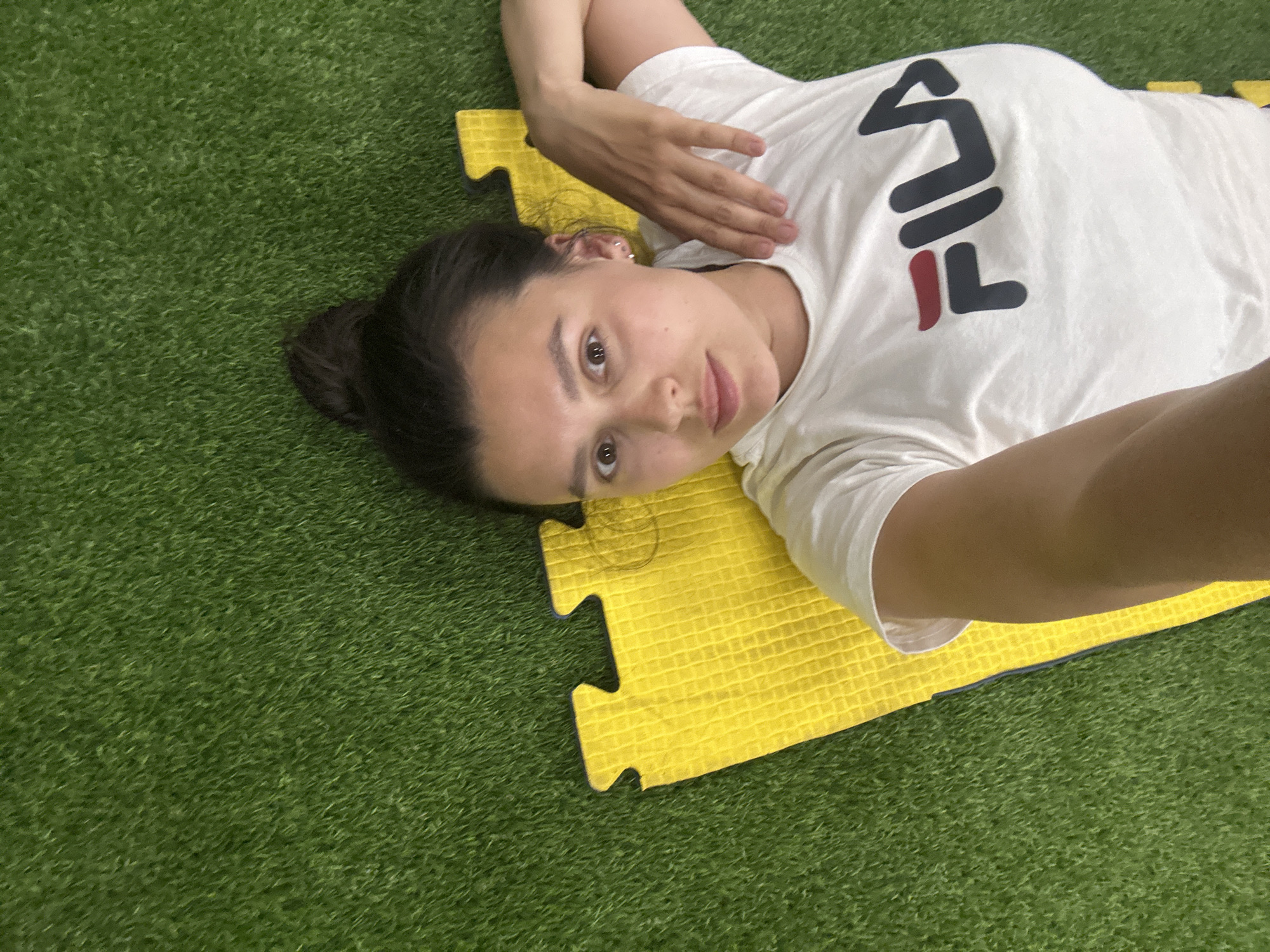 A woman is lying on a yellow exercise mat placed on artificial green grass. She is wearing a white t-shirt with a "FILA" logo. Her dark hair is pulled back in a bun. She is looking at the camera with a neutral expression. The scene is brightly lit. There is no indication of a specific location in Chaikovsky, Russia.