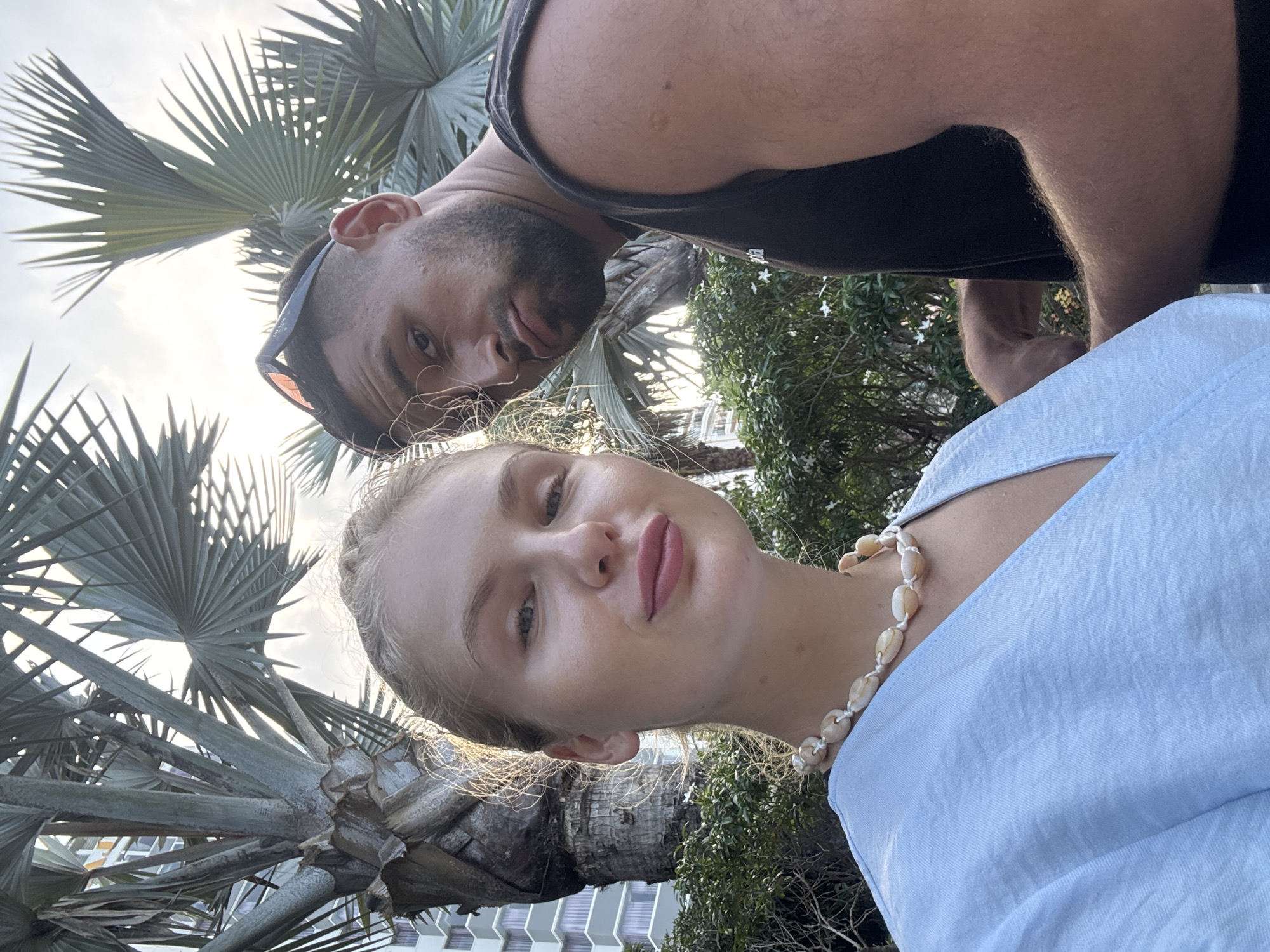 In Nha Trang Ward, Vietnam, a young Caucasian woman and a man of mixed ethnicity are captured in a selfie. The woman, positioned to the left, faces the camera with a slight smile and her hair pulled back. She wears a light blue top and a seashell necklace. To her right, the man is partially visible, looking towards the camera with sunglasses perched on his head and a beard. They are outdoors, with lush green foliage and palm trees forming the backdrop. A tall, modern building is discernible behind the foliage, suggesting an urban or resort setting. The lighting indicates it is daytime, possibly late afternoon or early morning, given the soft natural light.