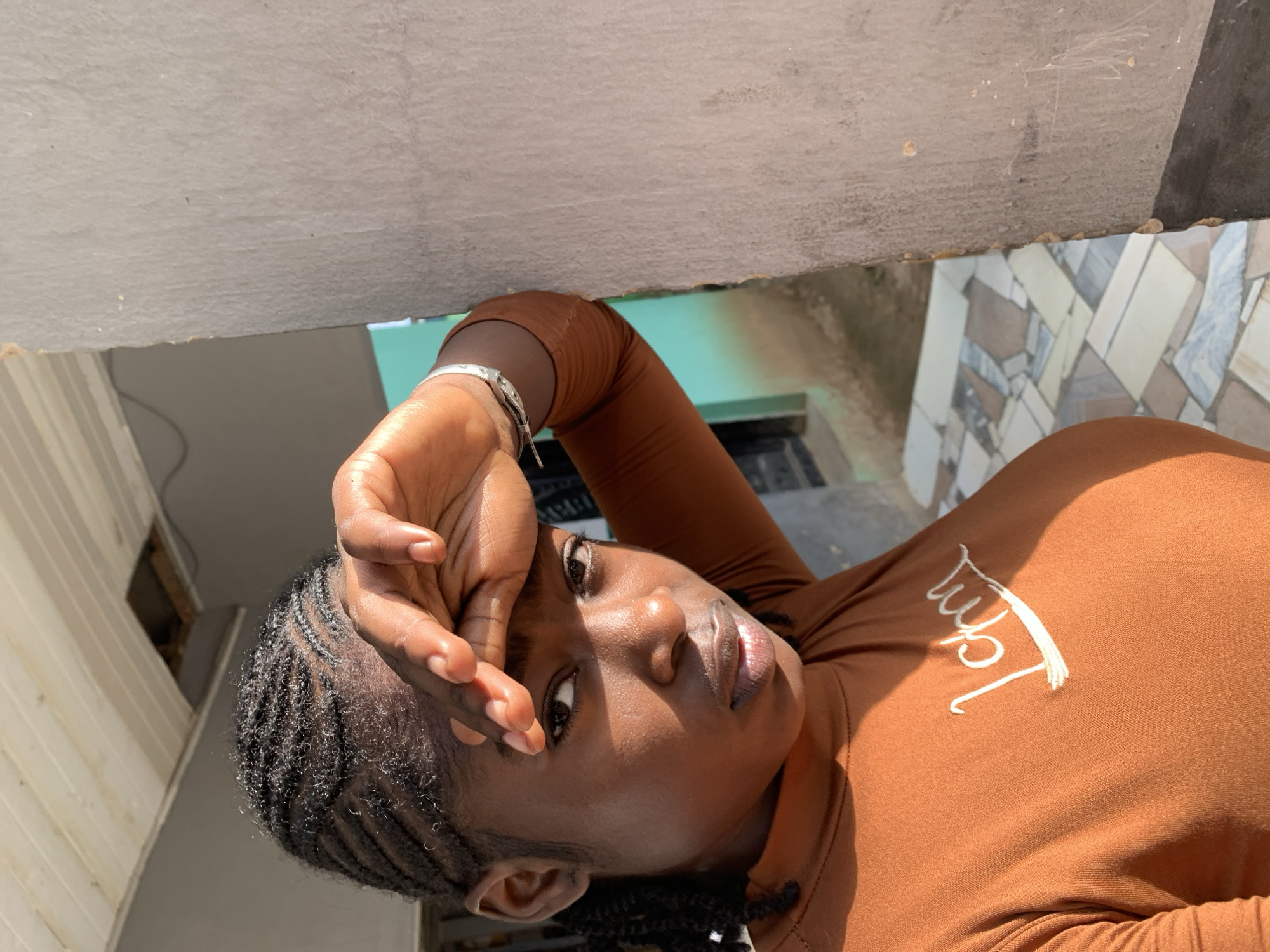 A young woman with dark skin and braided hair is depicted outdoors in the City of New Stadium, Nigeria. She wears an orange-brown long-sleeved top with white embroidered text, possibly "I'm", on the chest. Her left hand is raised to her forehead, fingers splayed, shielding her eyes from direct sunlight. A silver-colored bracelet is present on her right wrist. She leans against a textured, light-colored concrete wall occupying the right side of the frame, which shows minor surface irregularities. The background includes a light teal-colored wall, a dark rectangular frame (likely a window or door), and a white slatted ceiling with a dark opening. The ground features a patterned tiled floor in shades of grey, beige, and white. Strong sunlight illuminates the woman's right side, casting highlights and shadows.