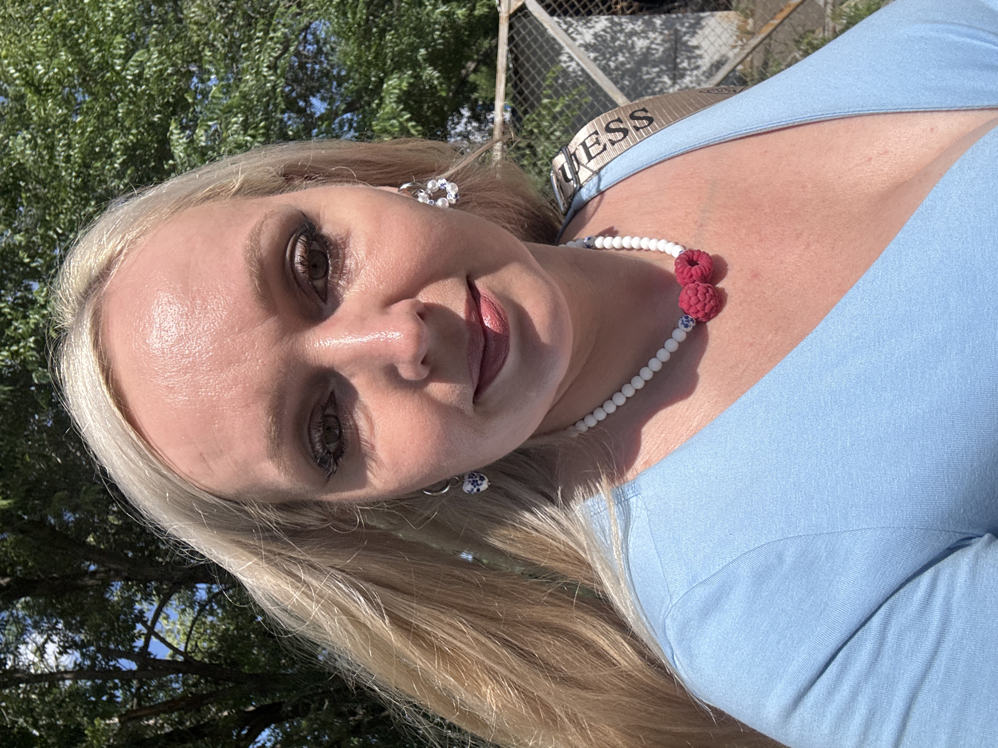 A blonde woman with fair skin and light makeup is shown from the chest up, facing the camera outdoors. She is wearing a light blue V-neck top, a white beaded necklace with raspberry-shaped charms, and hoop earrings with blue and white patterned charms. Her hair is long and blonde, falling over her left shoulder. Behind her, there are green trees and a chain-link fence. The lighting suggests it is daytime, with sunlight casting shadows. The location is not specified in the image itself, therefore, a specific location within the city of Komsomolsk-on-Amur, Russia cannot be determined from the visual information provided.