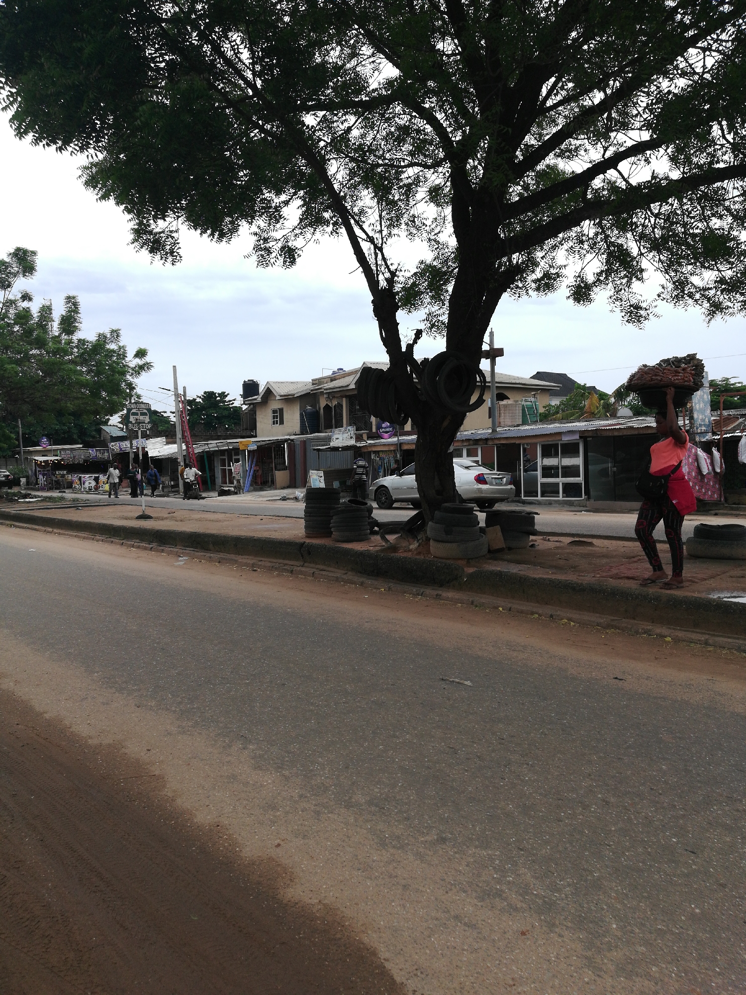 The image depicts an urban scene in Lagos, Nigeria. A tree with old tires hanging from it stands prominently on the roadside. A woman is walking along the sidewalk, carrying a load on her head and a red bag on her shoulder. Several small storefronts line the street behind the sidewalk, and a white car is parked nearby. Other people are walking in the background. The road appears to be paved, and the sky is overcast. A bus stop sign is visible in the distance.