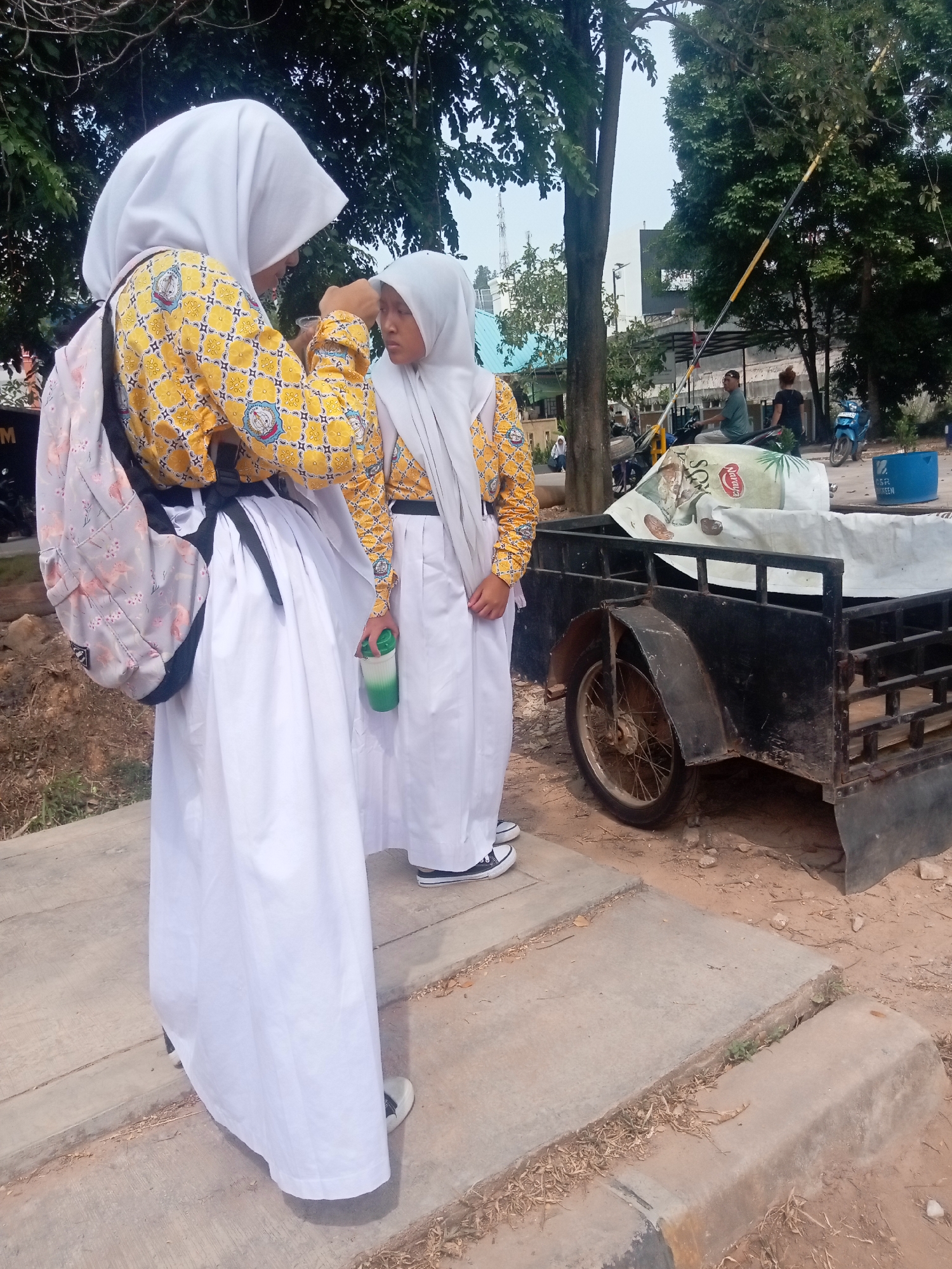 Two young females wearing hijabs and white long skirts, with yellow patterned tops, stand on a concrete pathway in an outdoor urban environment. The female on the left has a backpack with a floral pattern. The female on the right holds a green and white cup. They appear to be interacting. In the background, a black trailer with a spoked wheel is visible. Behind the trailer, several motorcycles and people are present, and a yellow and black barrier arm is raised. Trees and some buildings are also in the background, suggesting a location in the city of Batam, Indonesia. The ground is a mix of dirt and dry leaves.